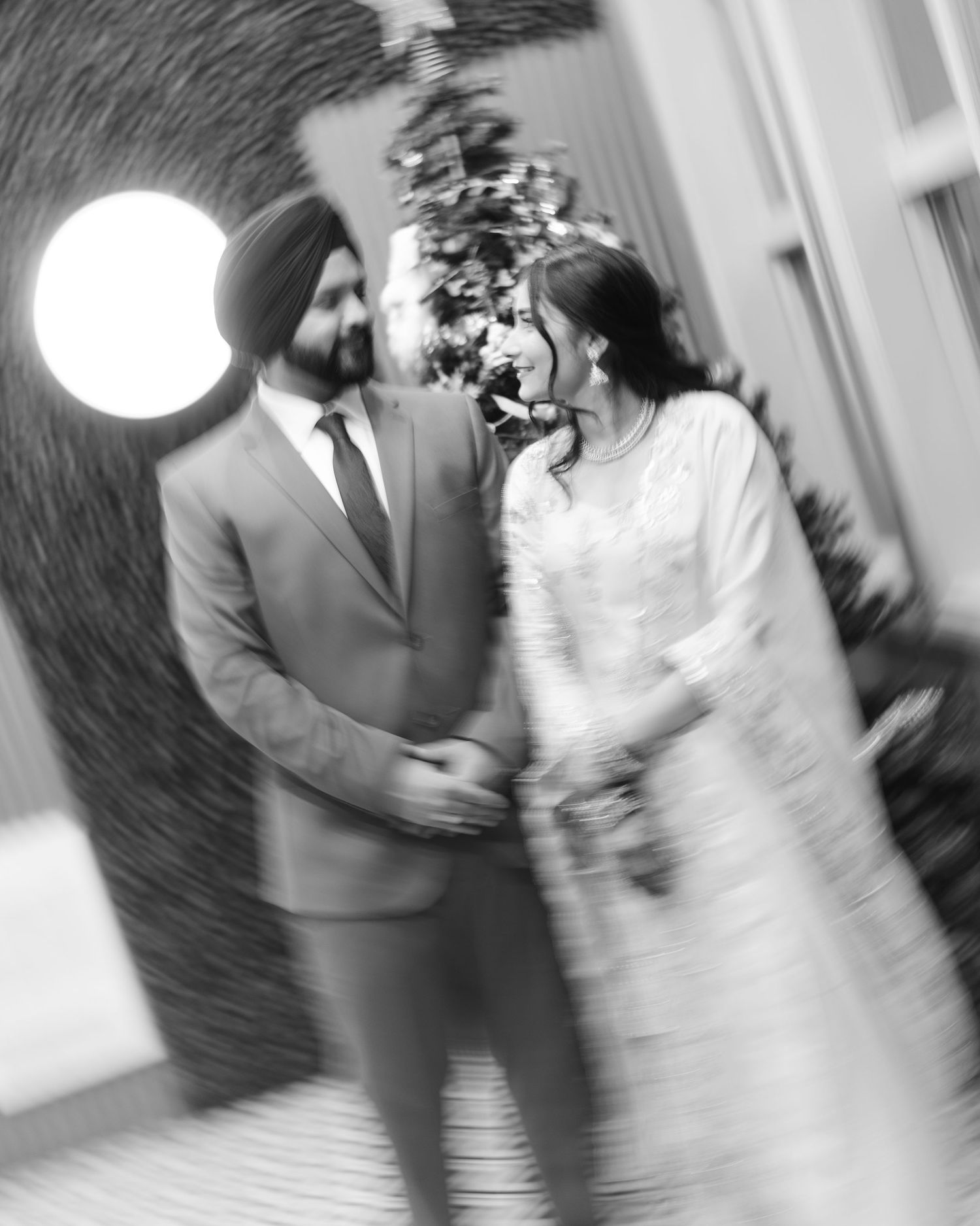 Black and white photo of Sikh couple holding hands and smiling at each other at elegant venue.