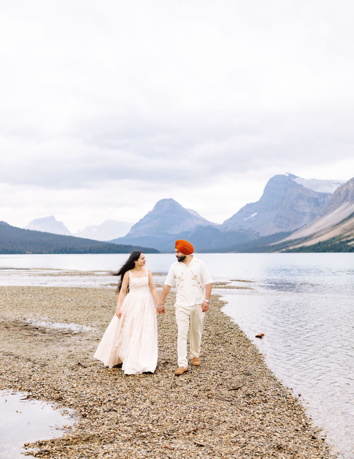 Couple in white attire strolling along scenic mountain lake shoreline under cloudy skies.