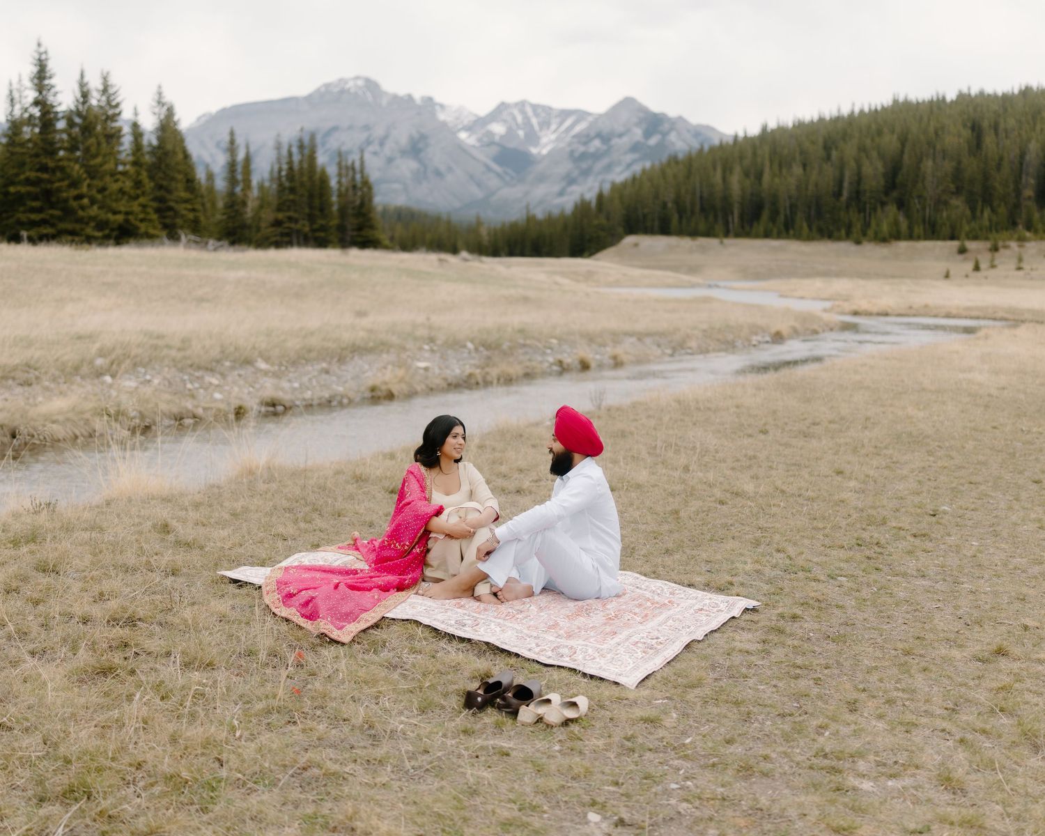 Sikh couple in traditional attire sitting on patterned blanket in open meadow with mountain backdrop.