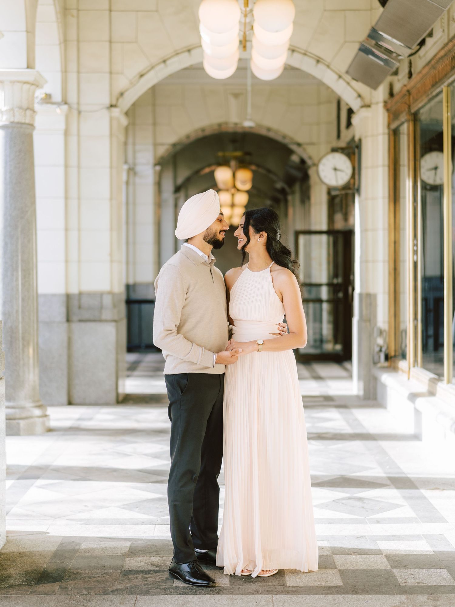 Sikh couple sharing intimate moment under ornate archway, woman in pleated blush halter gown.