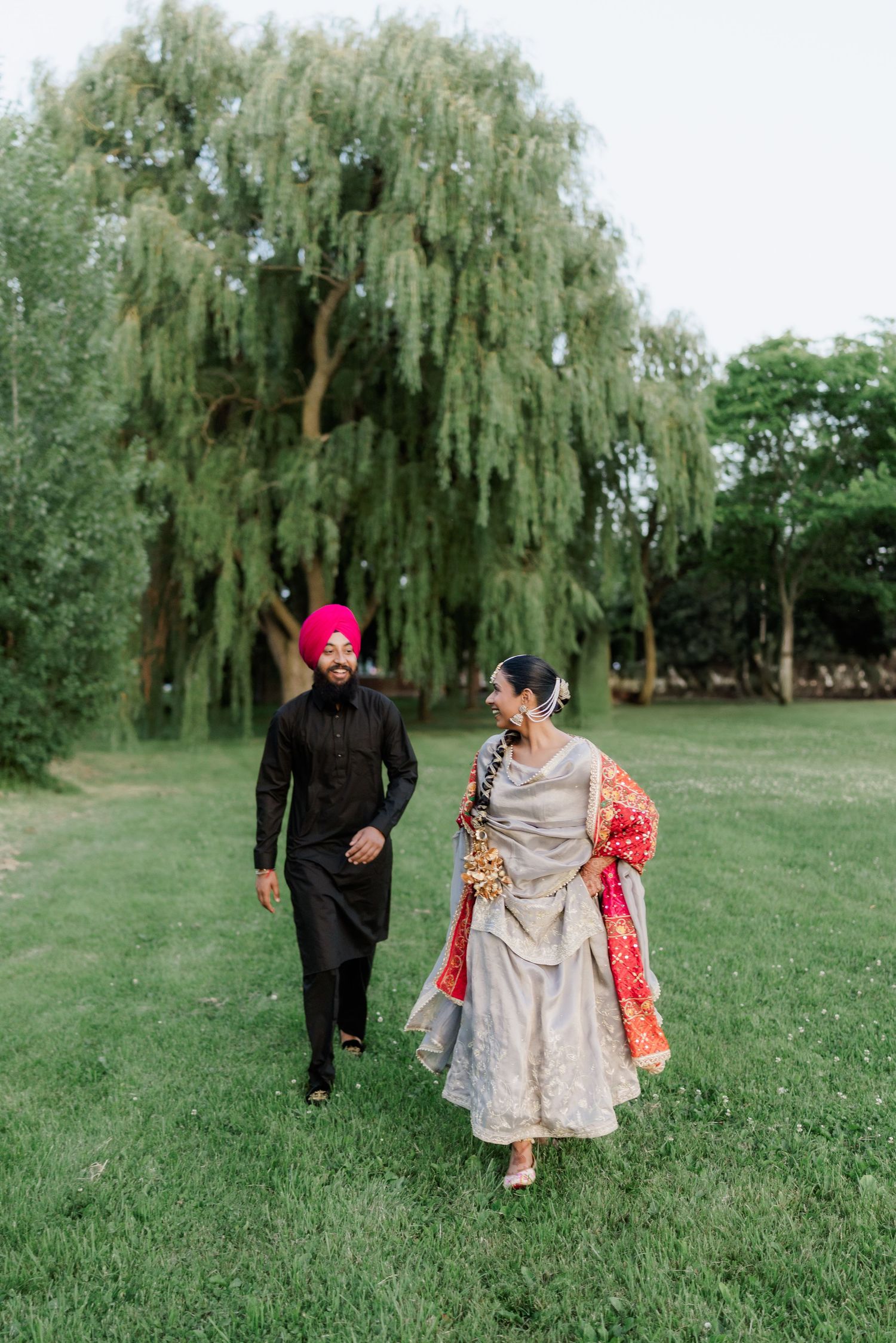 Sikh couple in traditional attire walking on grass beneath a weeping willow tree during outdoor celebration.