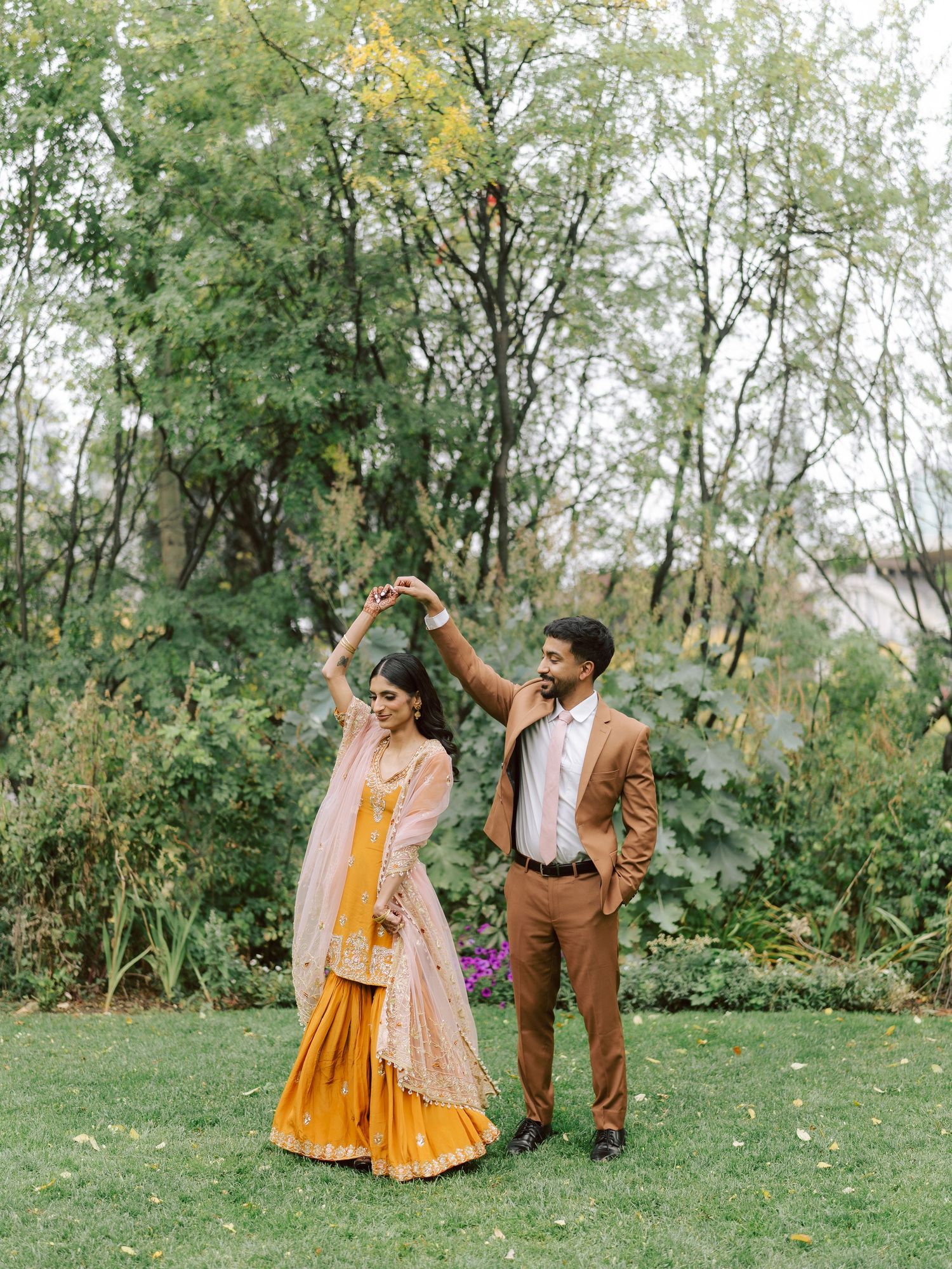 South Asian groom spins bride wearing yellow sharara in garden during outdoor wedding celebration.