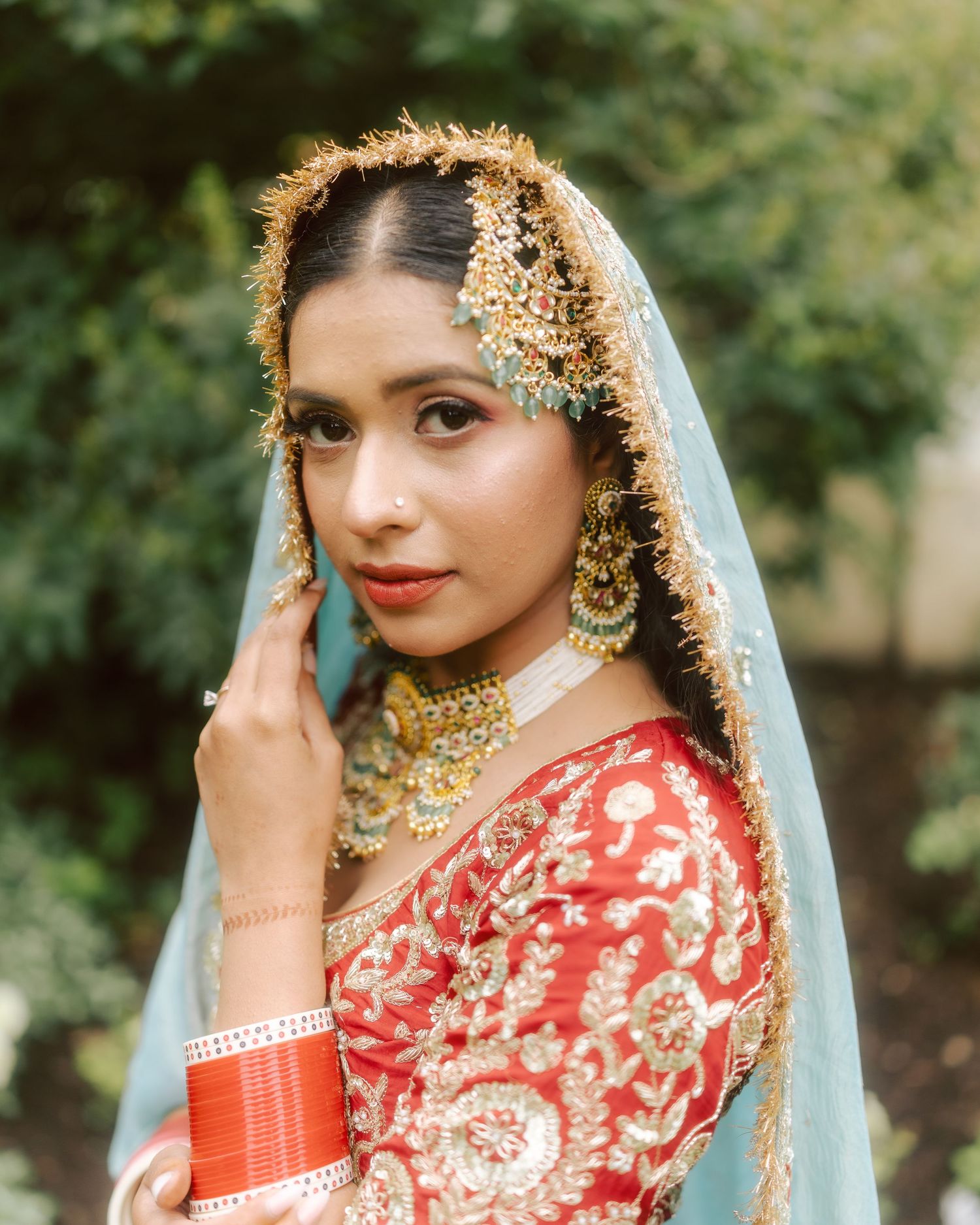 Close portrait of Sikh bride wearing gold jewelry maang tikka and light blue dupatta in garden.