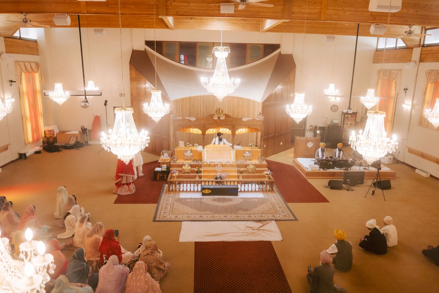 Interior of a Sikh Gurdwara with chandeliers, worshippers seated on floor, and ornate altar during religious ceremony.