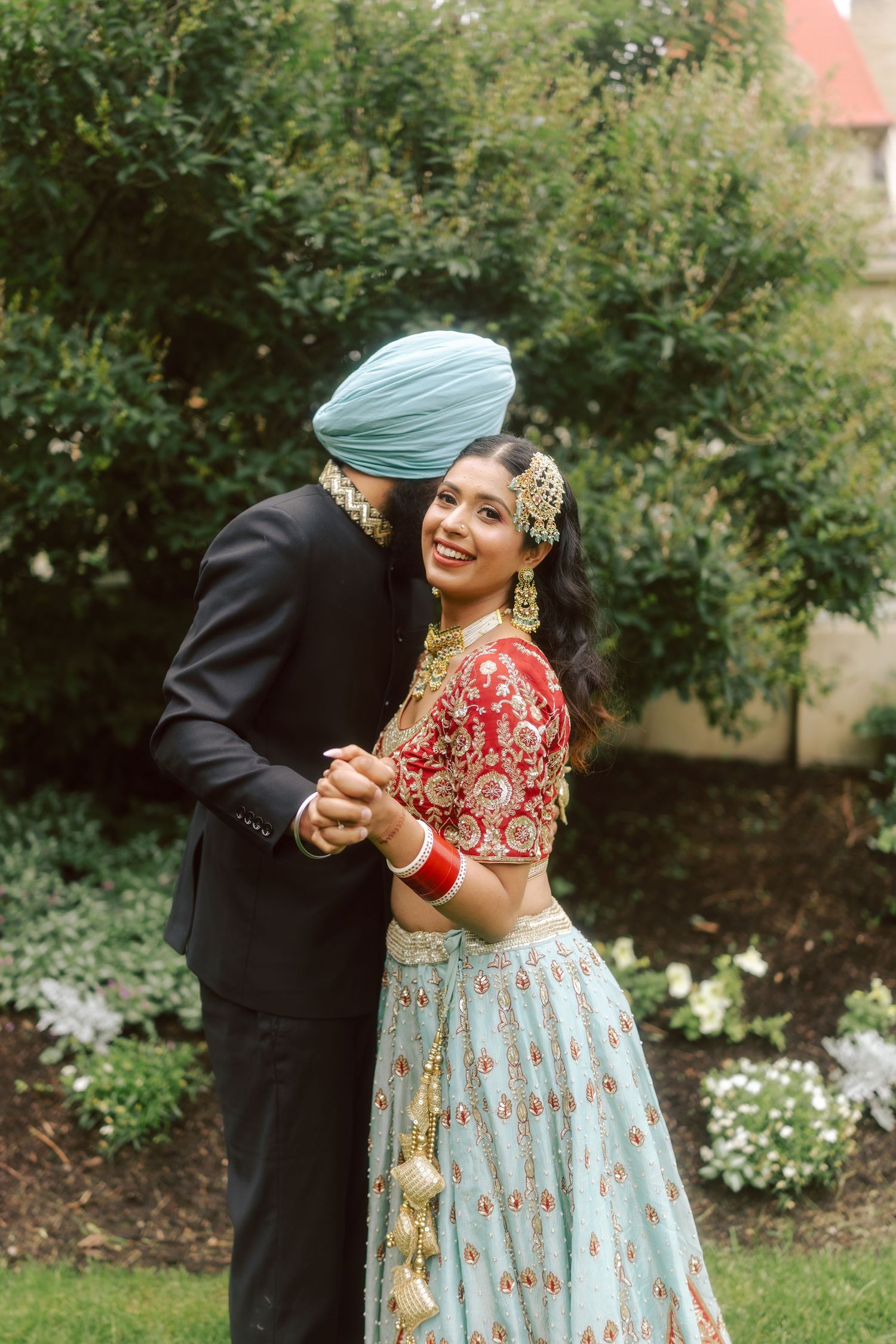 Sikh groom in blue turban kisses bride in red lehenga in garden setting.