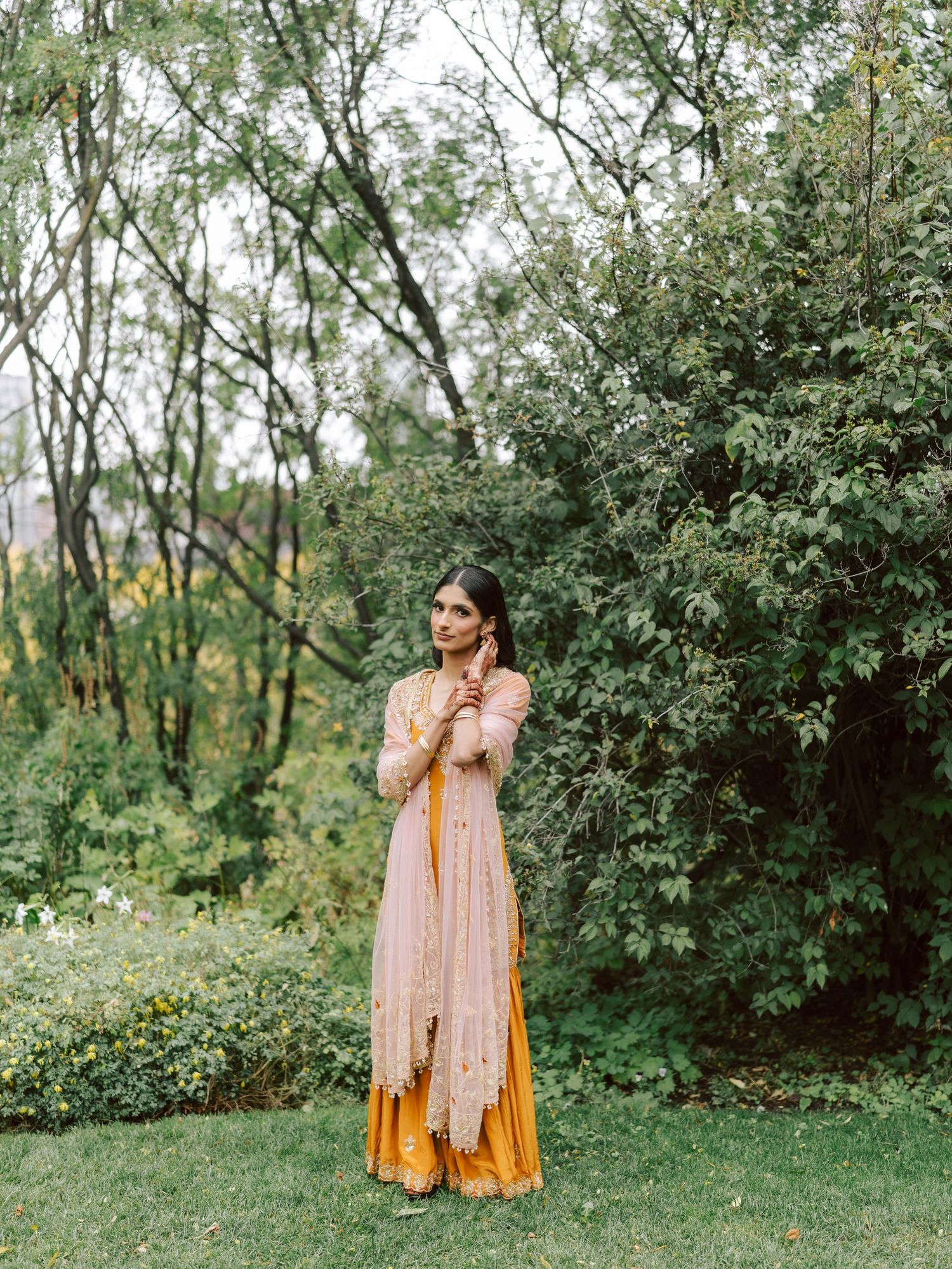 South Asian bride in mustard yellow and blush pink lehenga stands gracefully in garden setting.