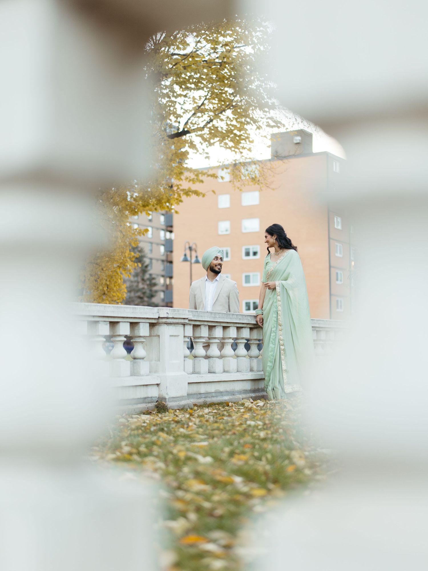 Couple in mint green traditional Indian attire standing by stone balustrade amid autumn foliage.