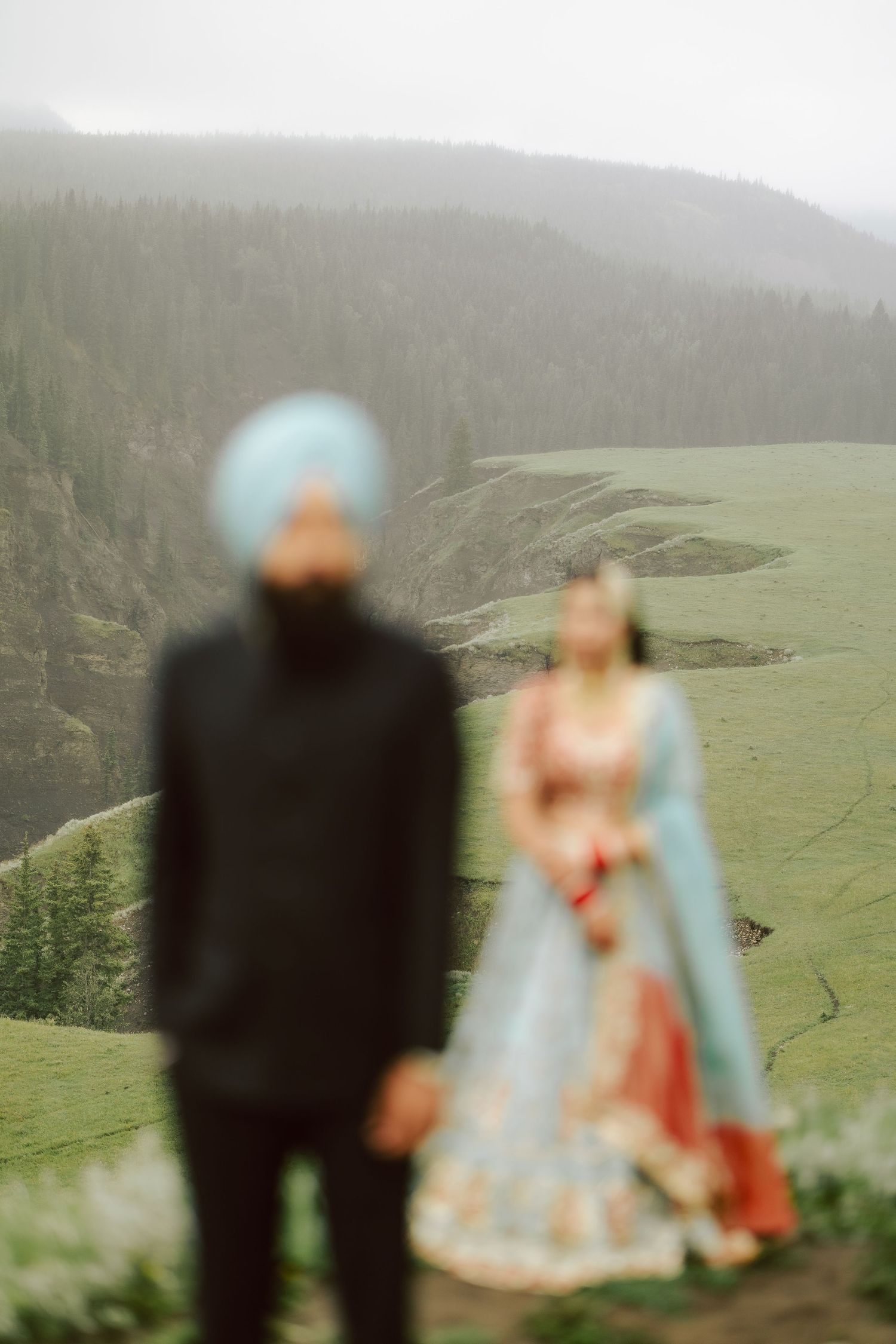 Defocused Sikh groom in blue turban with bride in red lehenga standing on scenic green hillside.
