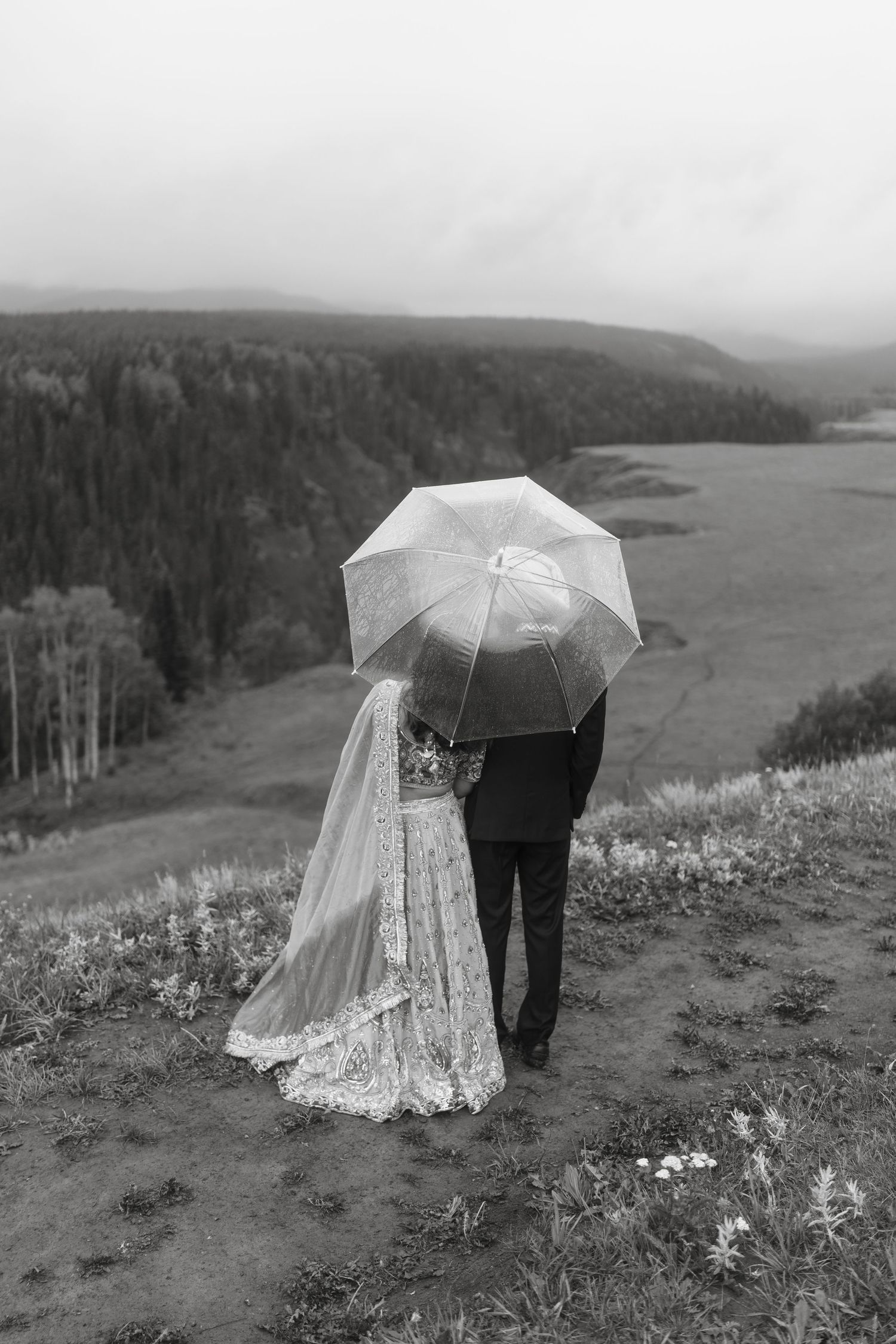 Black and white photo of bride and groom sharing umbrella overlooking misty valley landscape.