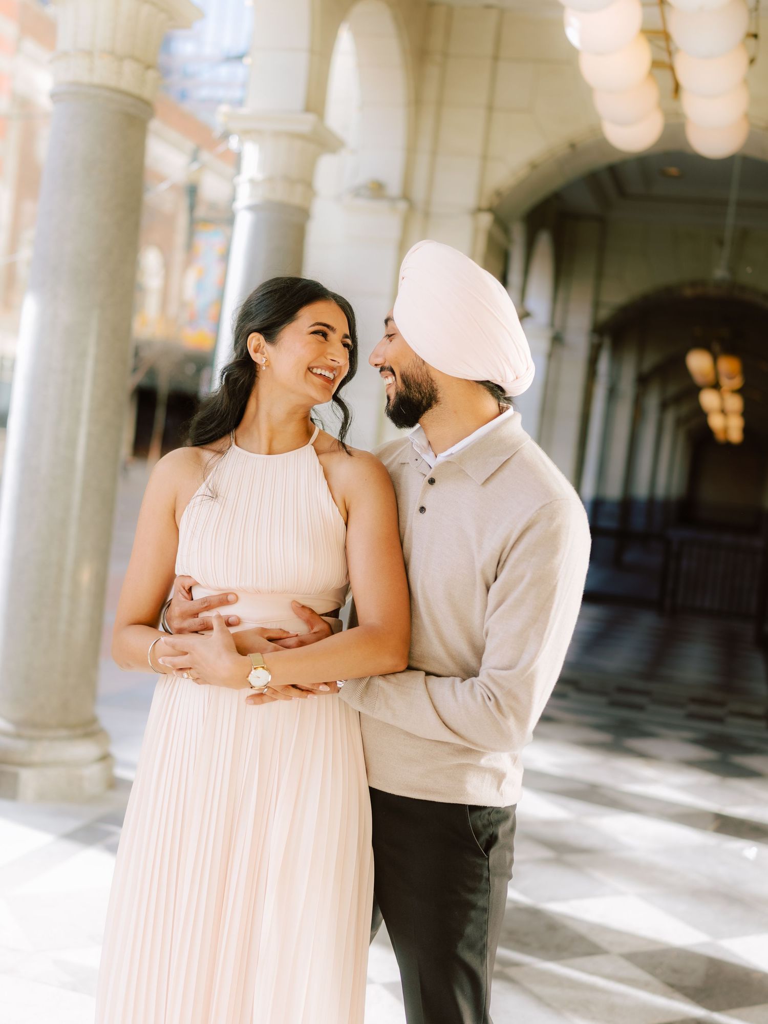 Joyful Sikh couple laughing together in elegant colonnade, woman in blush pleated halter dress.