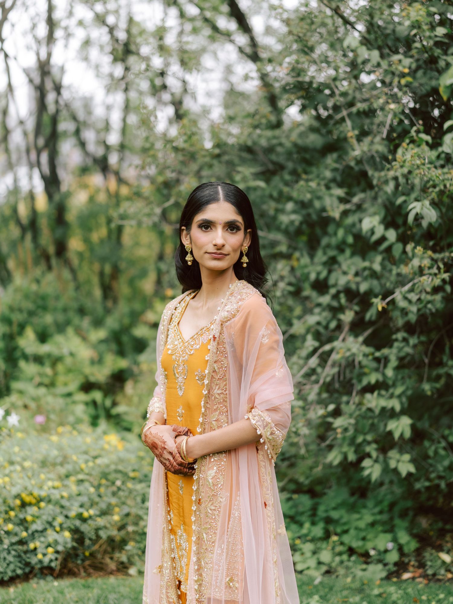 South Asian bride in yellow embroidered sharara with blush dupatta poses gracefully in garden setting.
