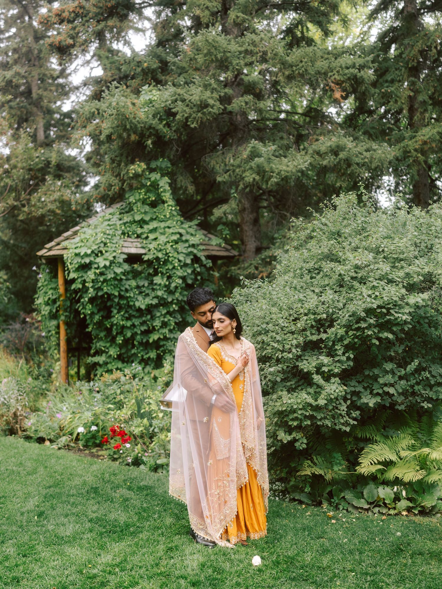South Asian couple in traditional wedding attire embrace in green garden with gazebo backdrop.