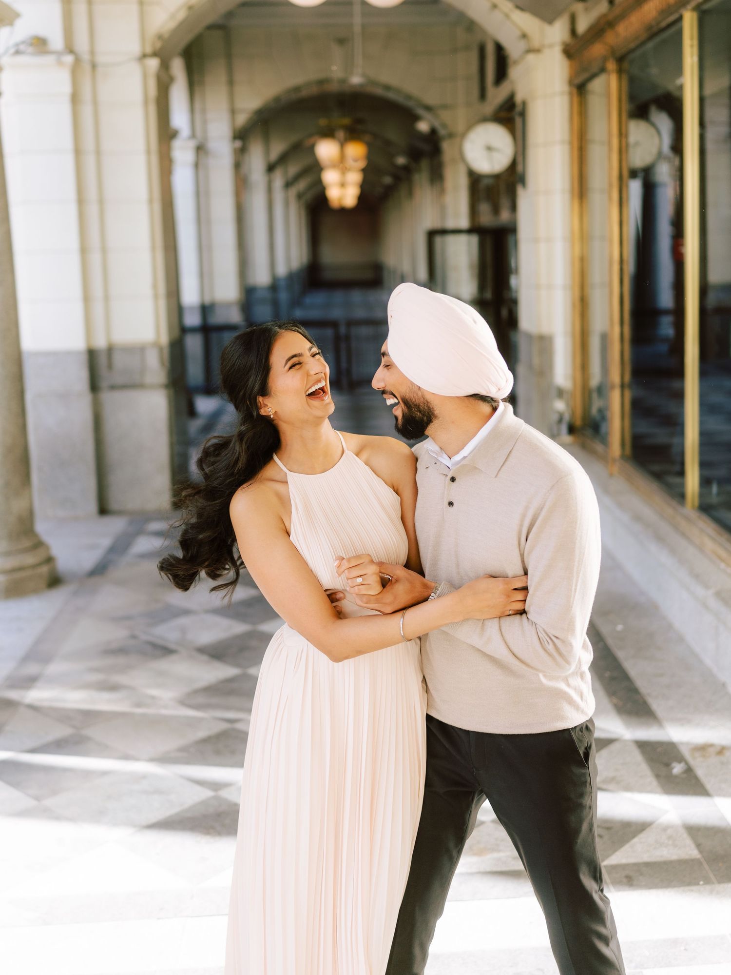 Laughing couple embracing in elegant arcade hallway, woman in white pleated dress, man in white sweater.