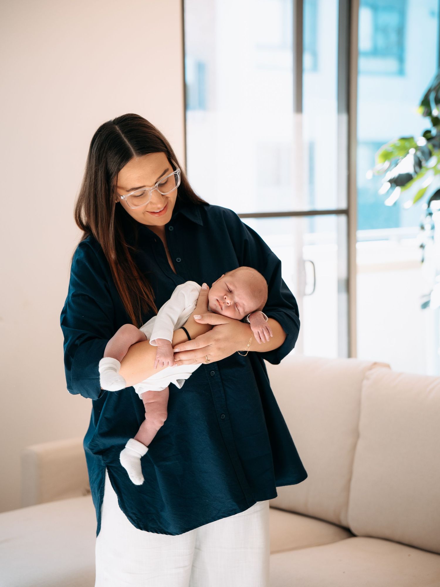 A woman in a dark blue shirt and glasses tenderly holds a sleeping newborn baby indoors.