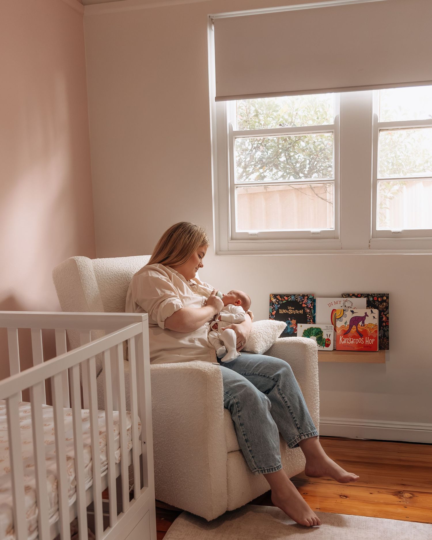 Mother breastfeeding newborn baby in white nursery glider chair beside white crib in pink room.