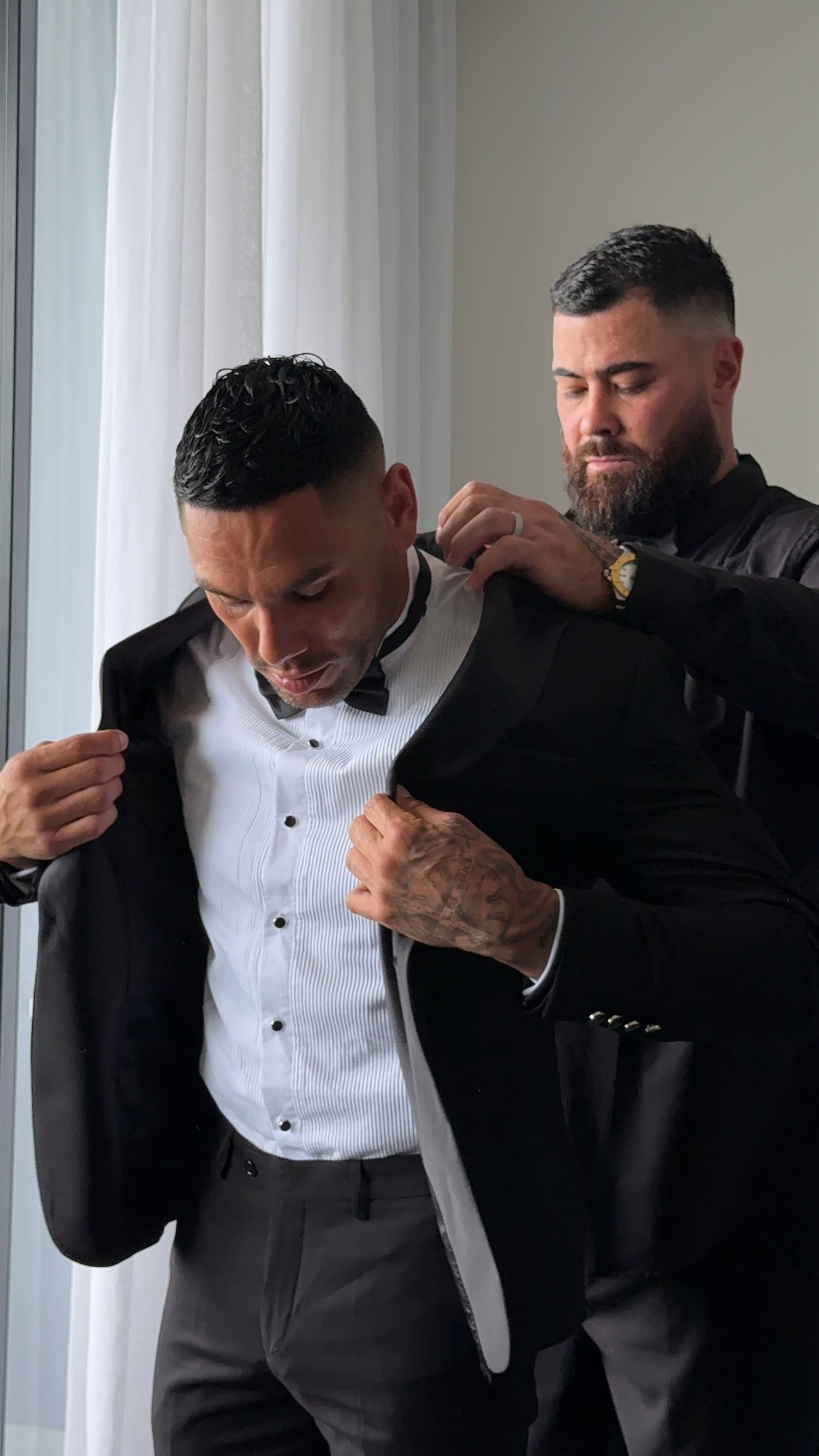 Two men in formal attire help each other get dressed, with one adjusting the other's suit jacket.