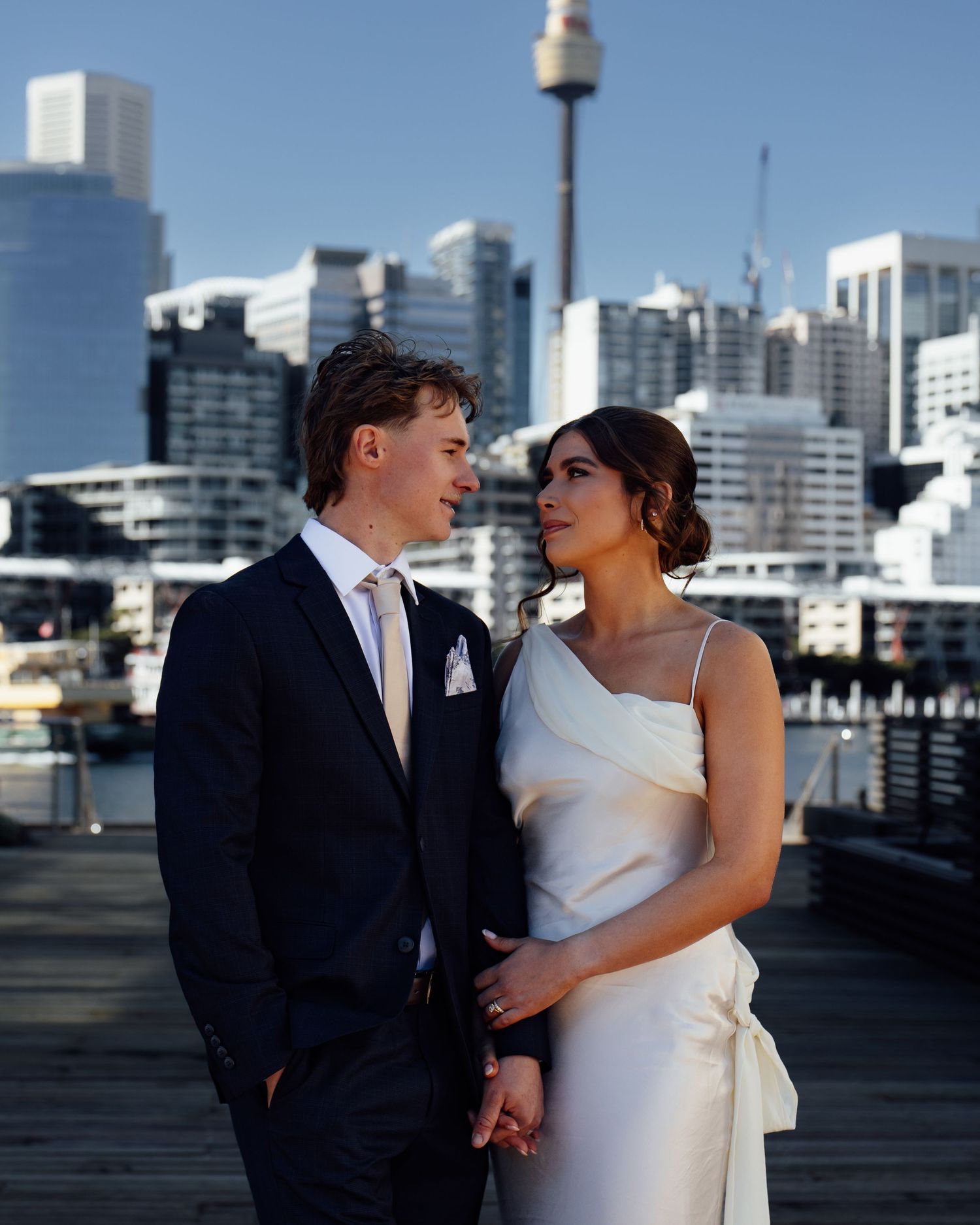 A bride and groom pose on a waterfront dock with Sydney city skyline and tower in the background.