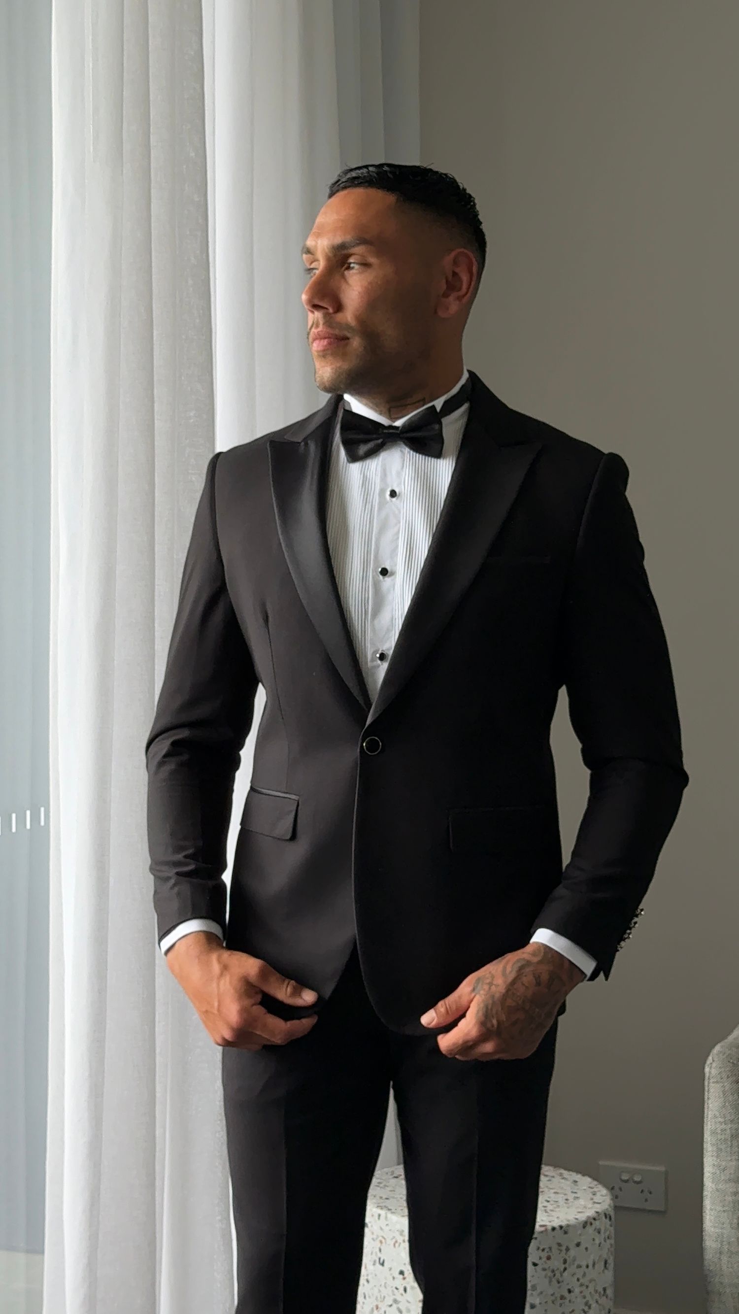 A person wearing a classic black tuxedo with bow tie stands by a window in elegant formal attire.
