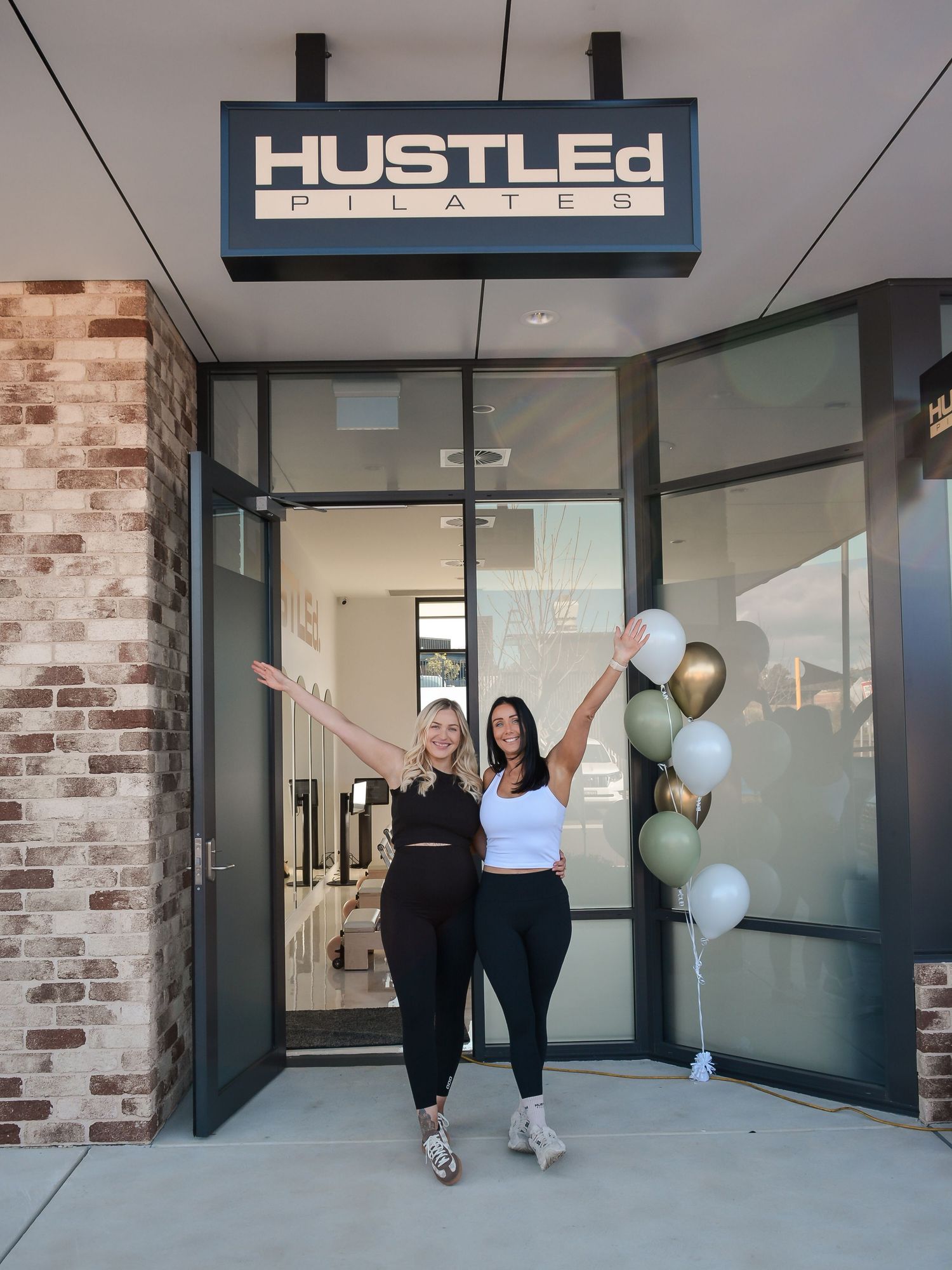 Two people celebrating with balloons outside a storefront with 'HUSTLED' signage.