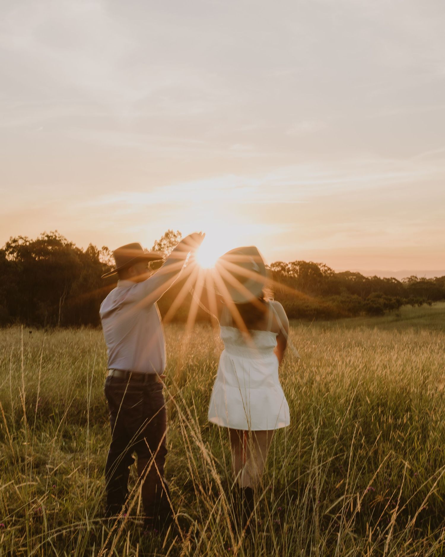 Couple dancing in golden sunset field, man in cowboy hat lifting woman's arm, sun flare glowing between them.