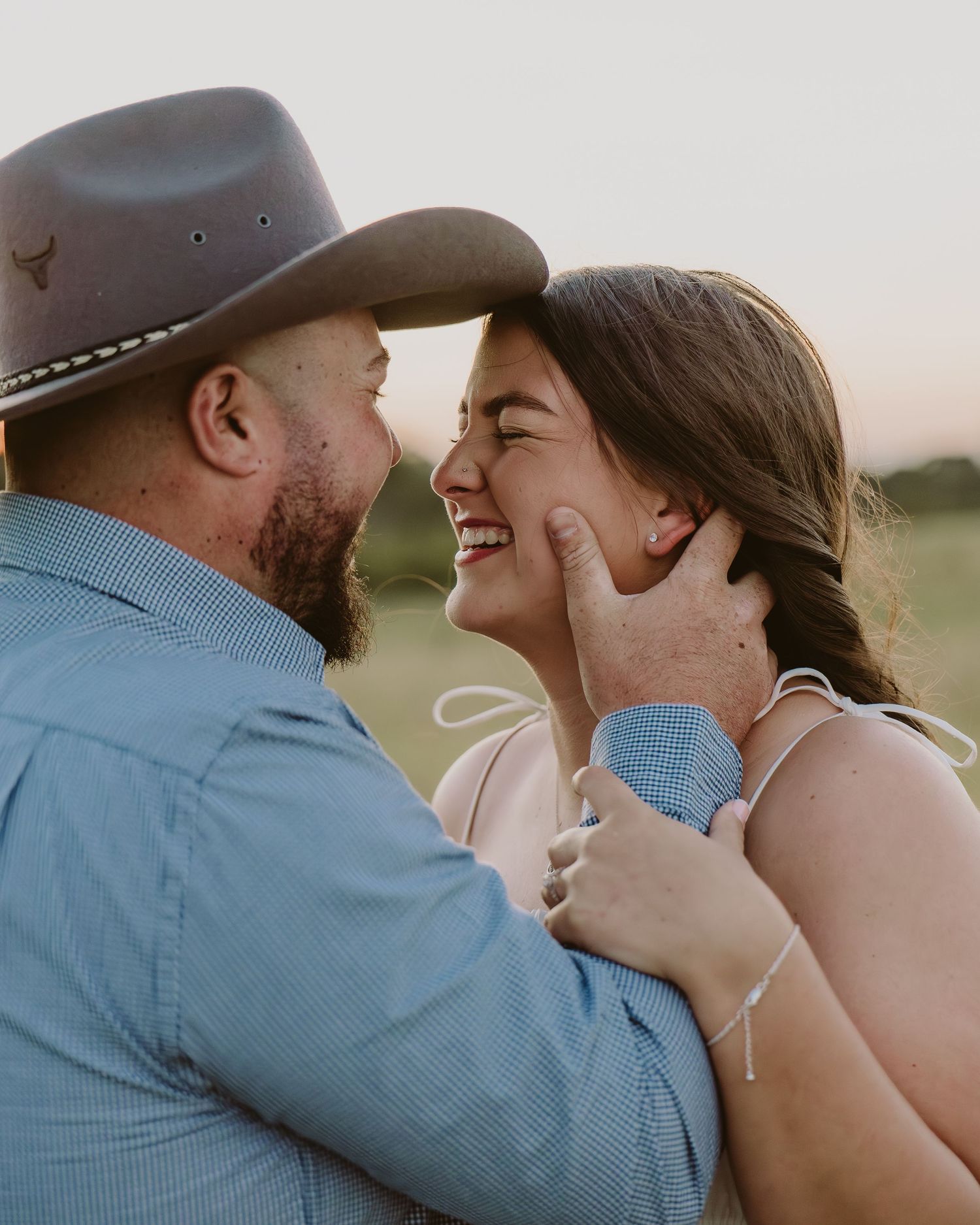 A couple shares a joyful moment outdoors, man in cowboy hat tenderly holding woman's face as she laughs.
