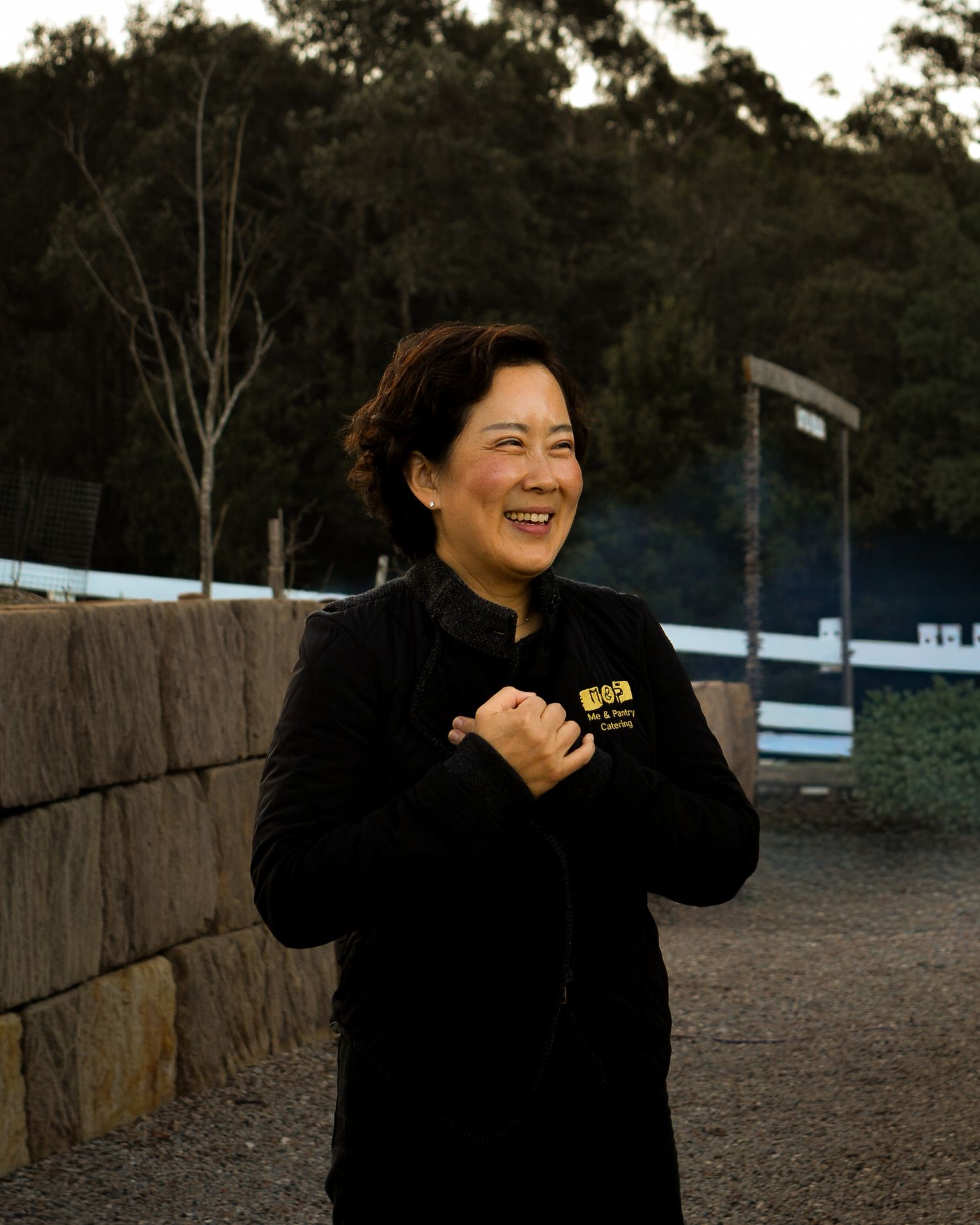 Person in black clothing laughing joyfully outdoors against a natural backdrop.