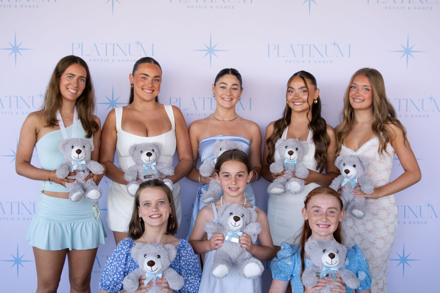 Group photo of dancers in light blue dresses holding teddy bears against a branded backdrop.