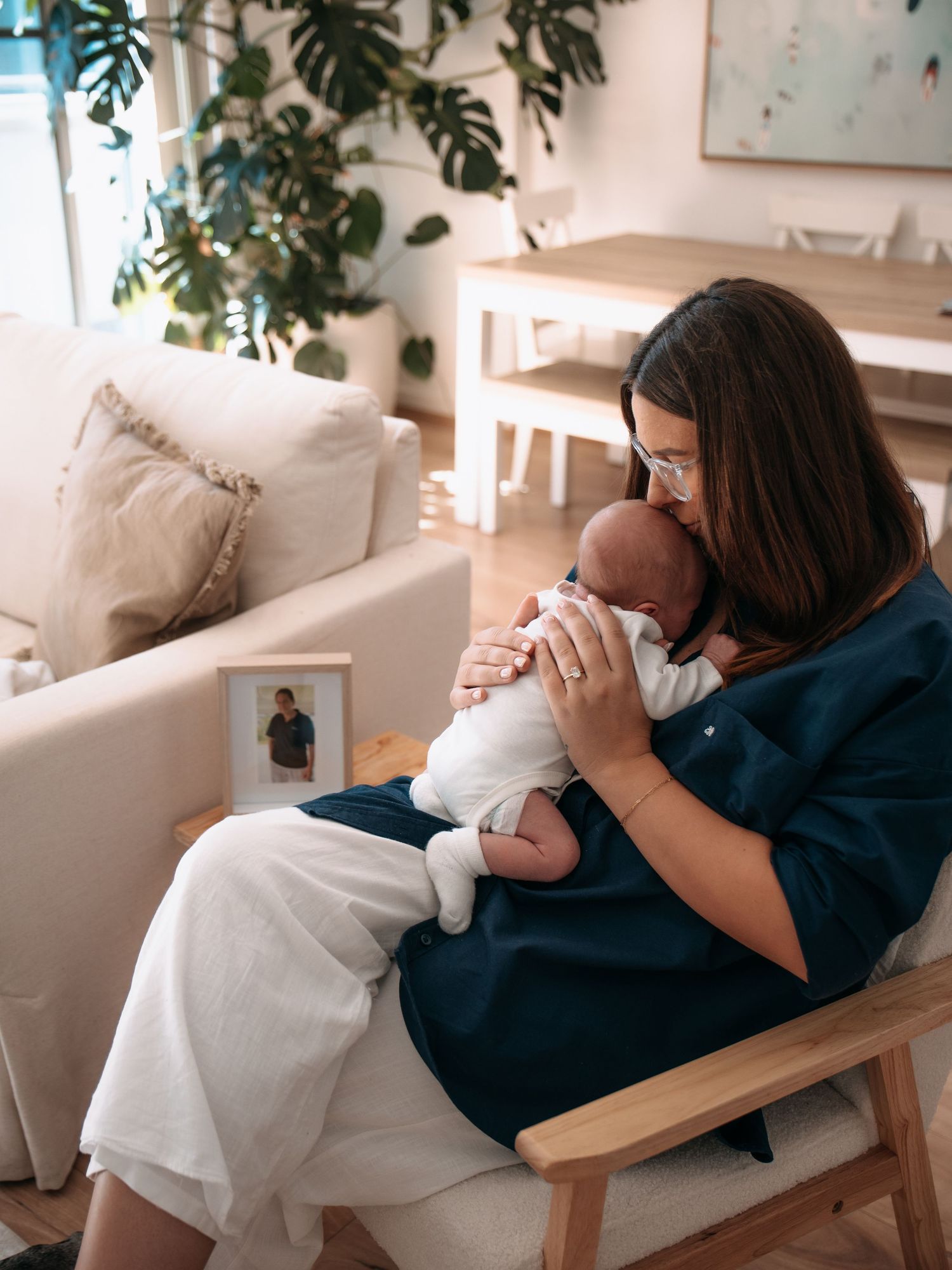 A mother in a teal top gently kisses her newborn baby while seated in a wooden chair in a cozy living room.