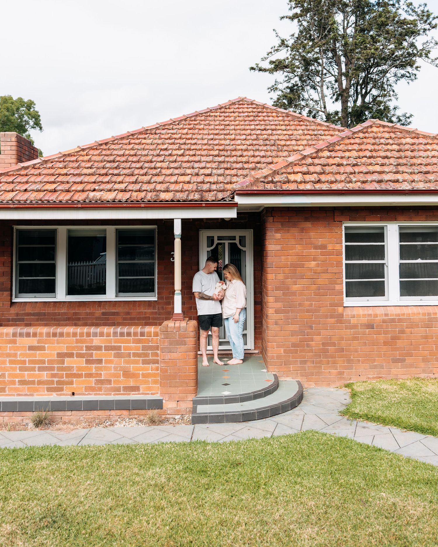 Couple standing at the front door of a red brick home with a tiled roof and green lawn.