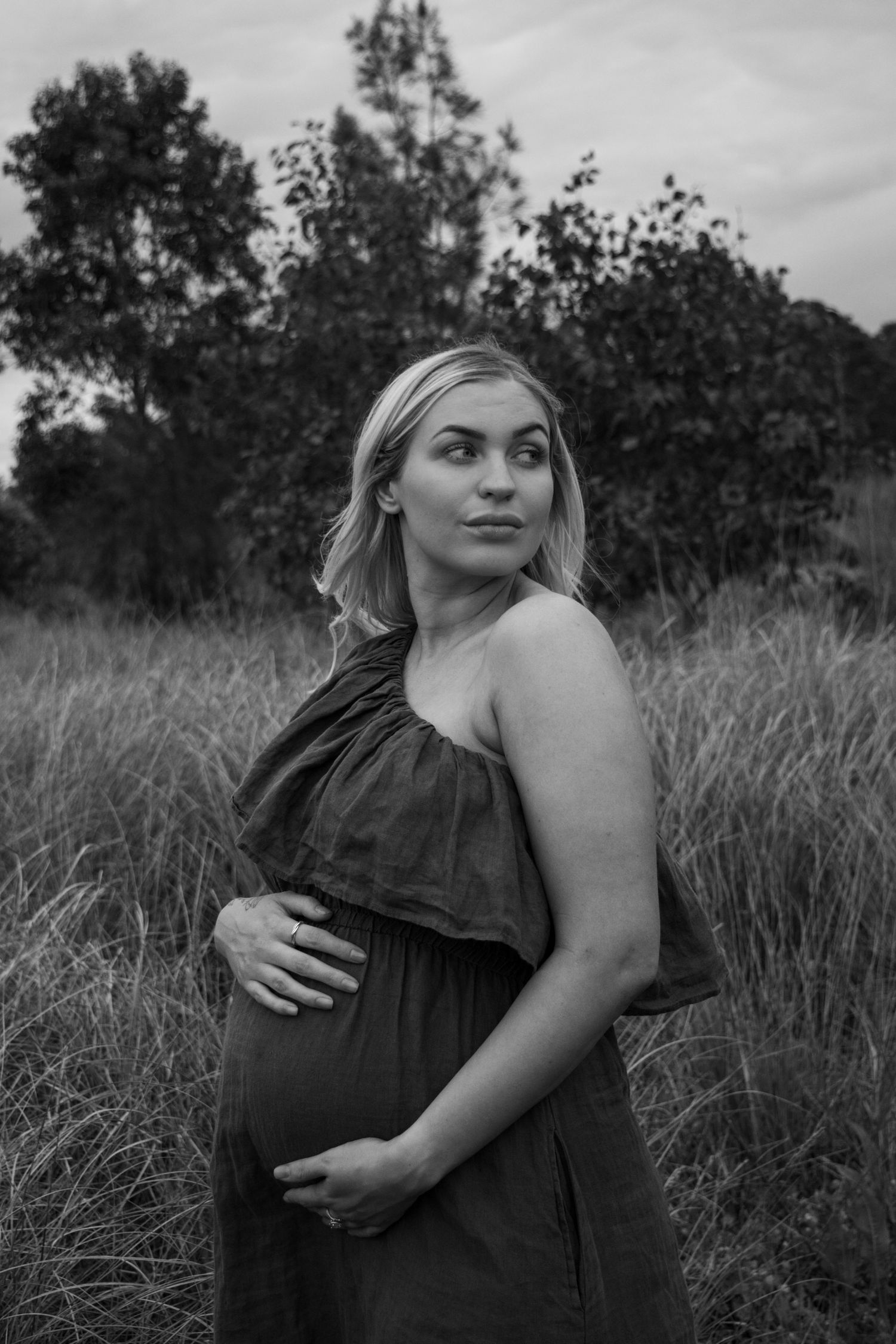 A black and white maternity portrait of an expectant mother in a flowing dress standing in a field of tall grass.
