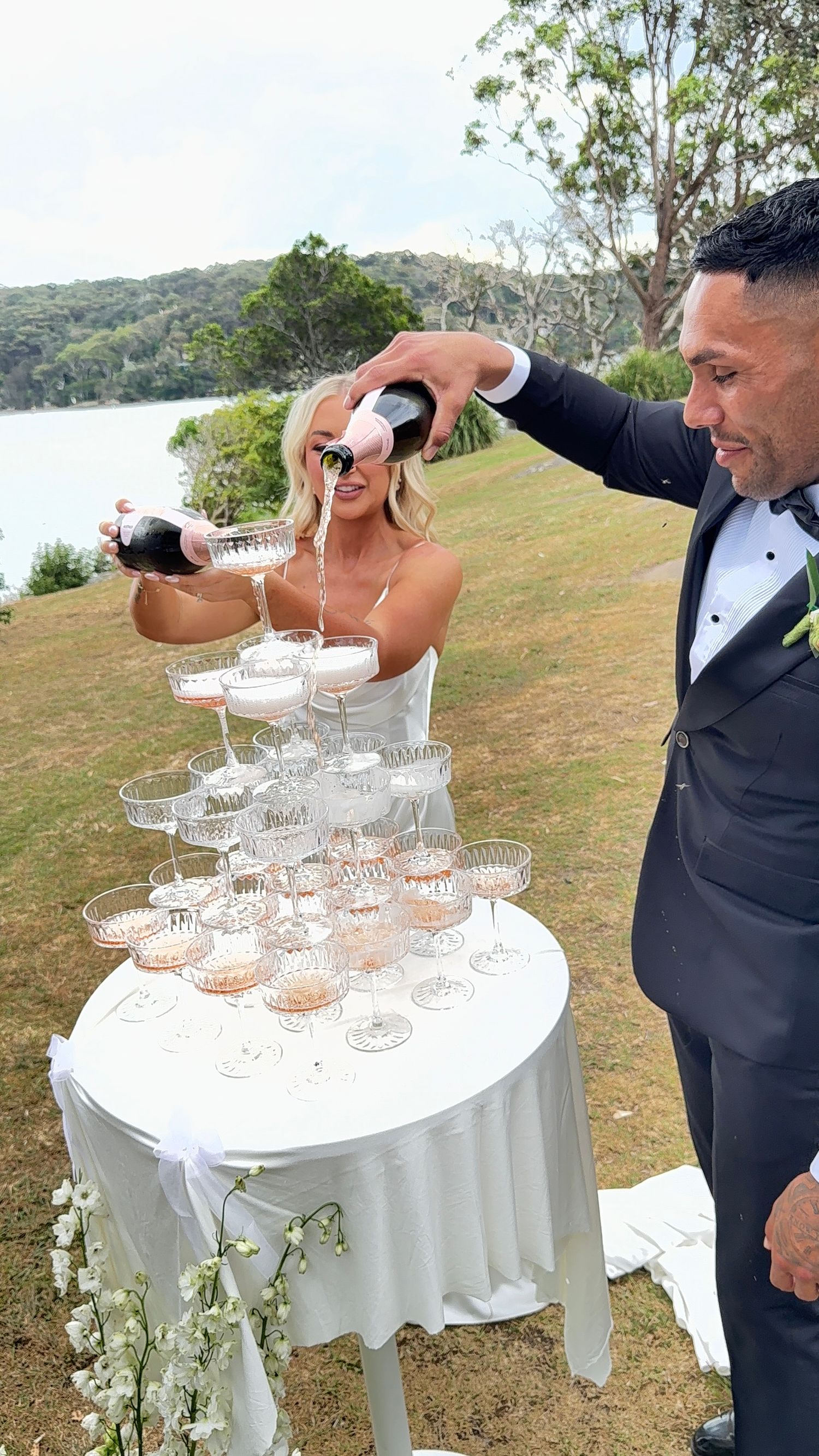 A wedding couple builds a champagne tower outdoors with crystal glasses on a white-clothed table near a scenic waterfront.