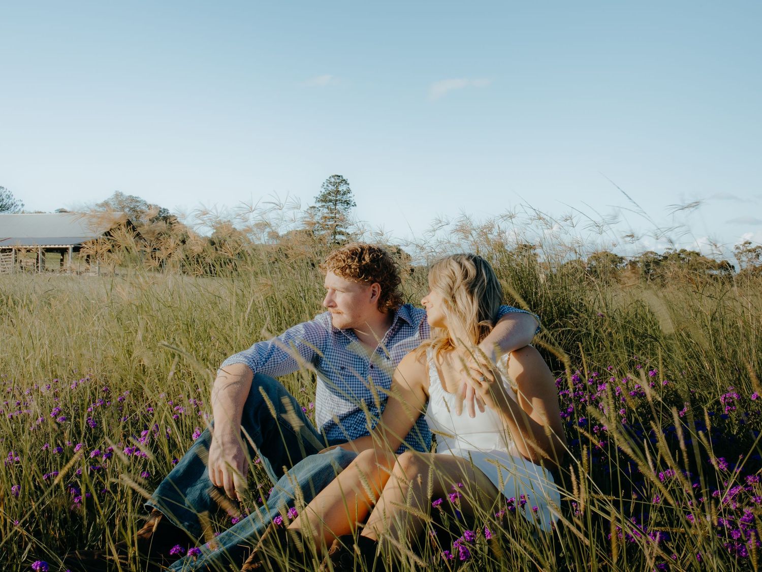 A couple sits romantically in a lush green field with purple wildflowers under a clear blue sky.