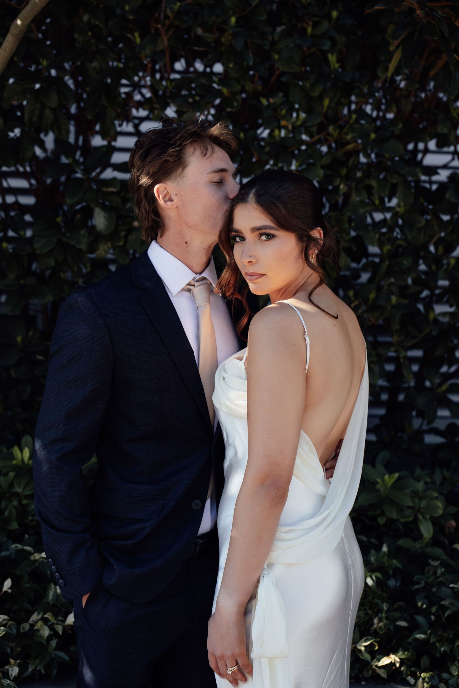Couple in wedding attire pose outdoors, groom in navy suit kisses bride wearing elegant white satin backless gown.