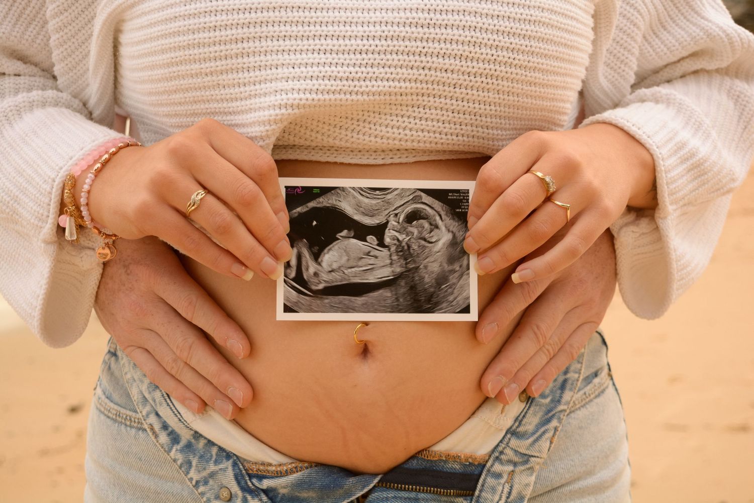Hands holding ultrasound photos against a pregnant belly in casual attire.