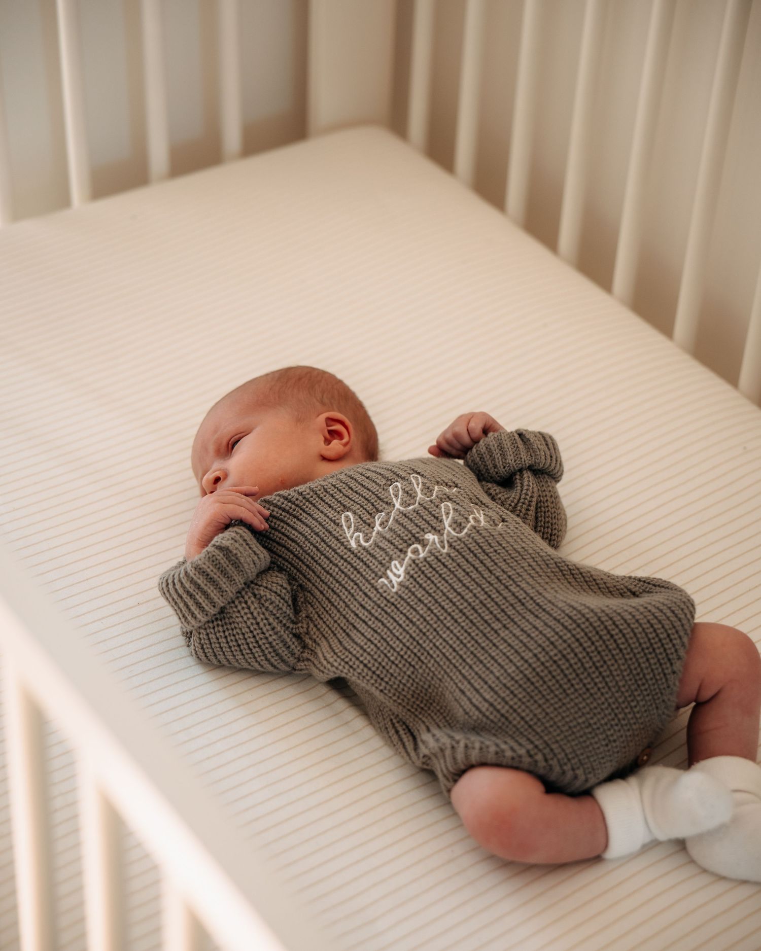 Newborn baby sleeping peacefully in a white crib, wearing an olive knit romper embroidered with hello world.