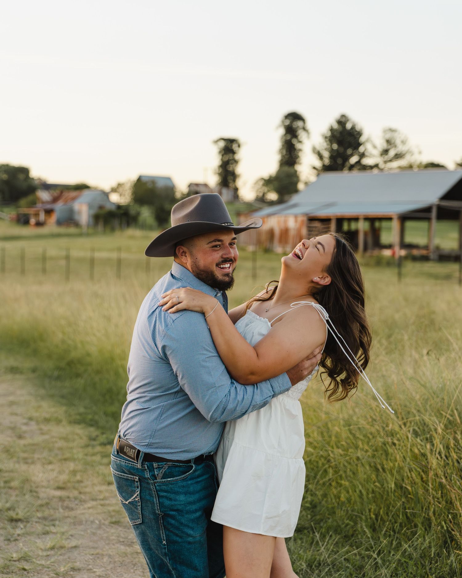 A joyful couple laughs and embraces in a green field at golden hour, man wearing cowboy hat and blue shirt.