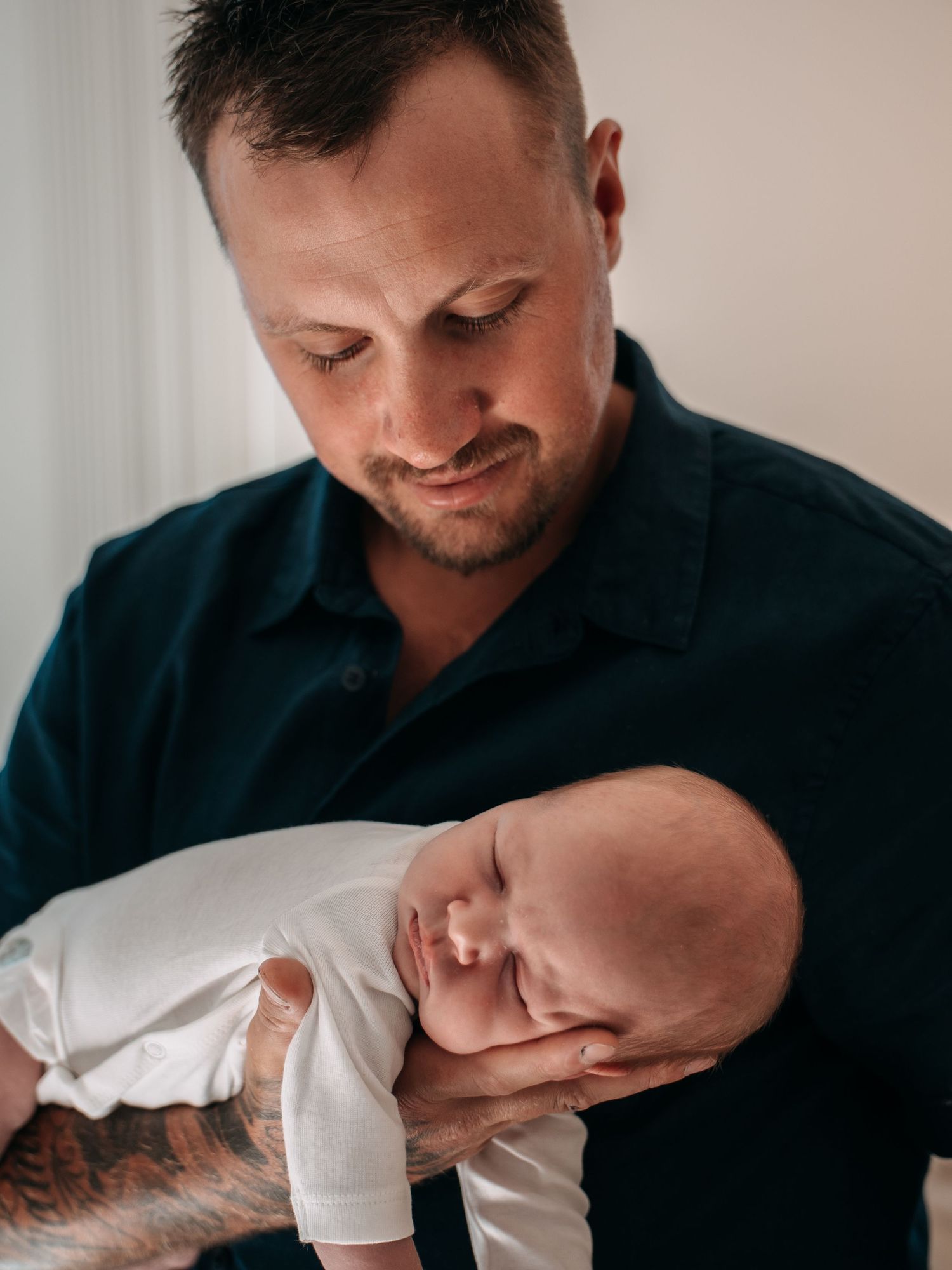 A tattooed father tenderly holds his sleeping newborn baby in his arms, gazing down lovingly.