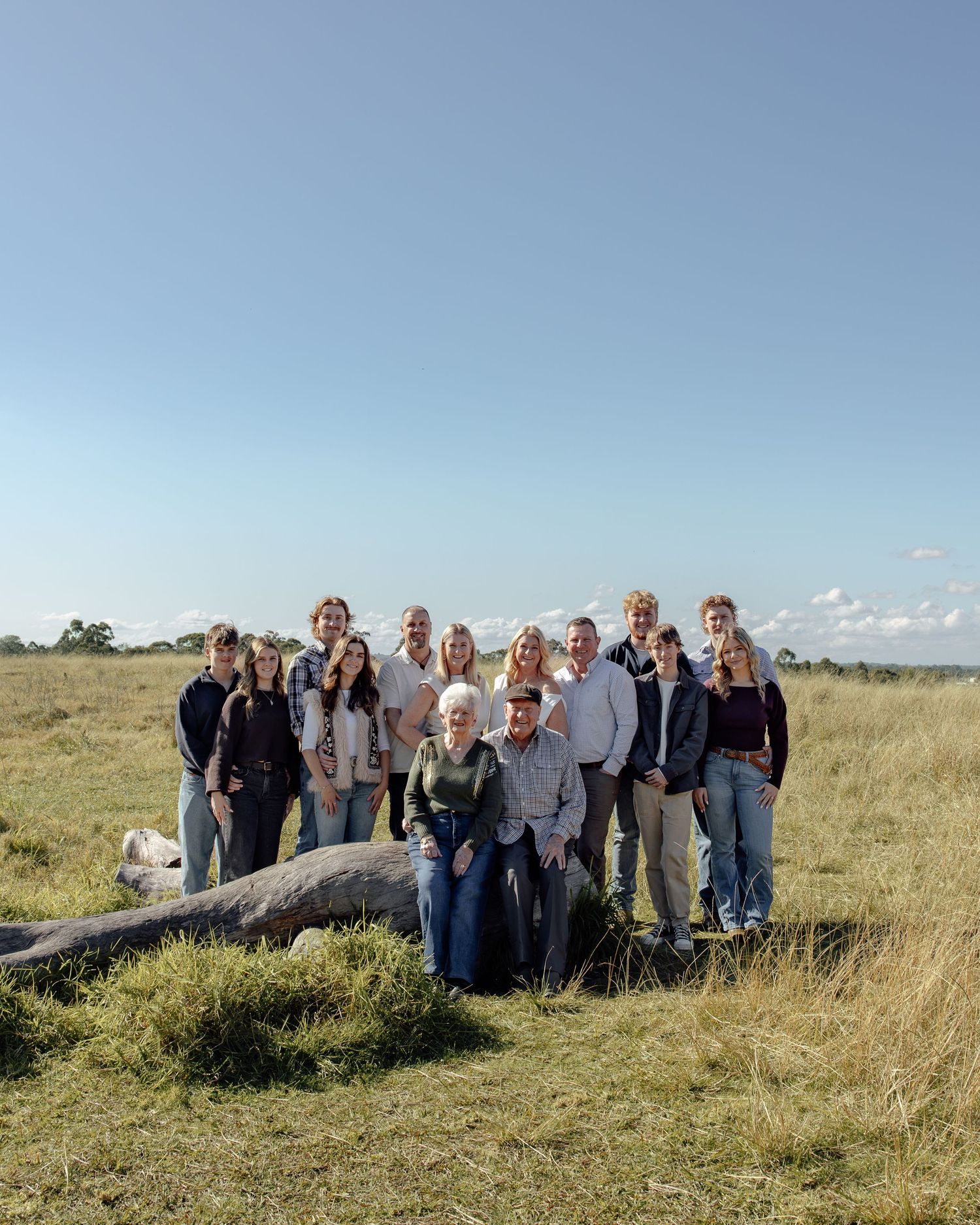 Large family group portrait outdoors in grassy field under blue sky, posing near a fallen log in a rural landscape.