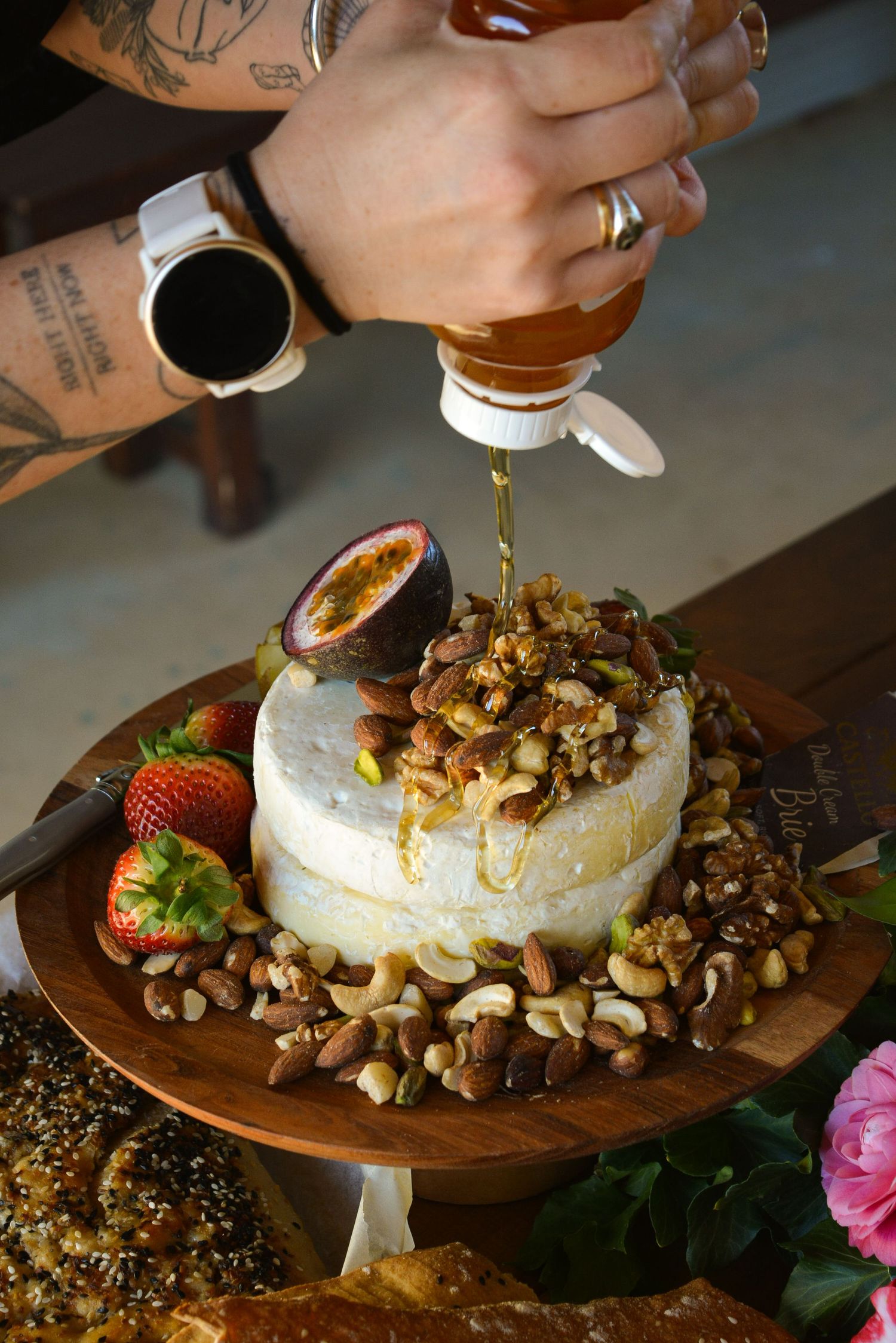 Hands drizzling honey over a baked brie topped with nuts on a wooden serving board.