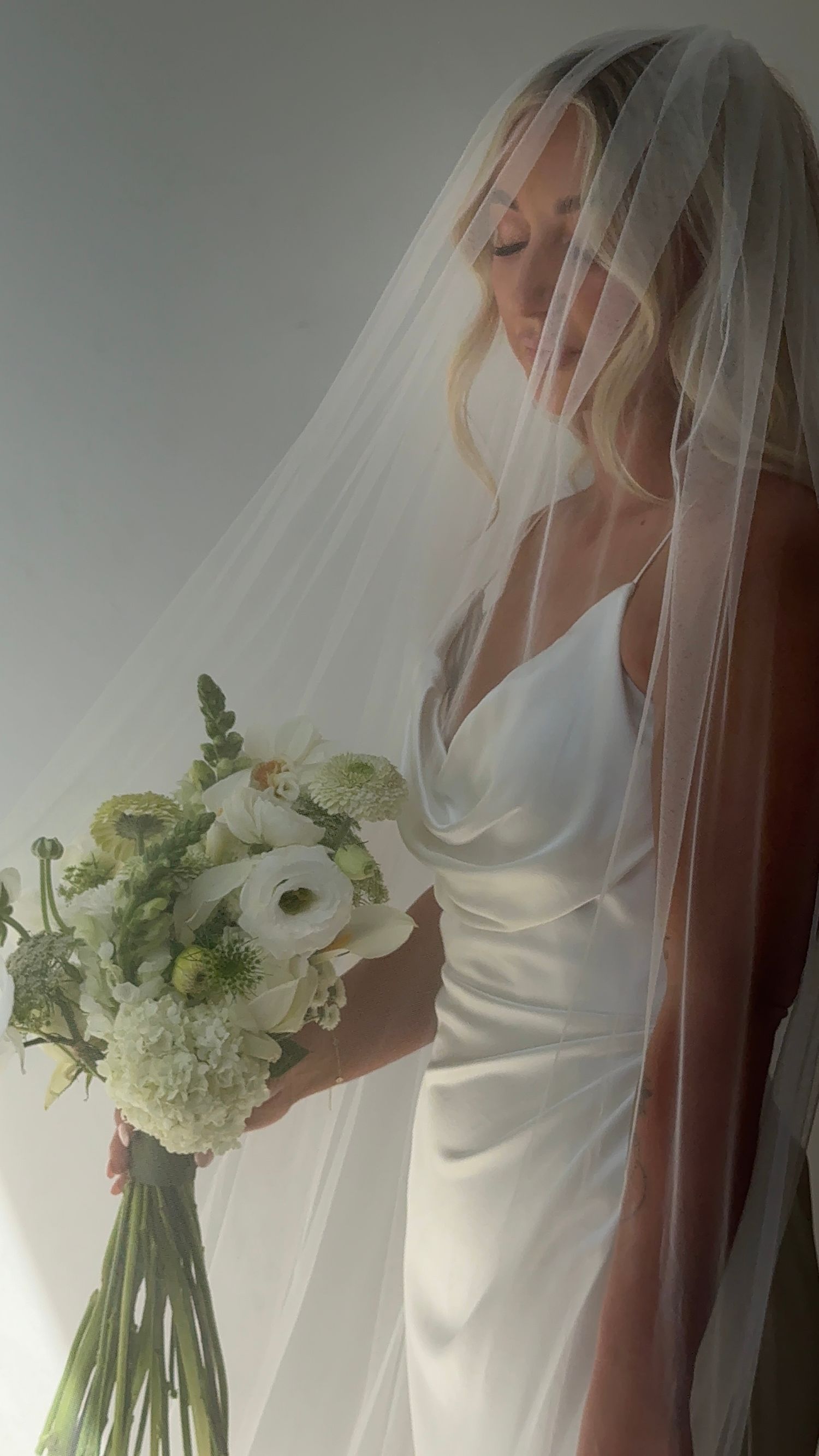A bride in a flowing white wedding dress and veil holds an elegant bouquet of white and green flowers against natural light.