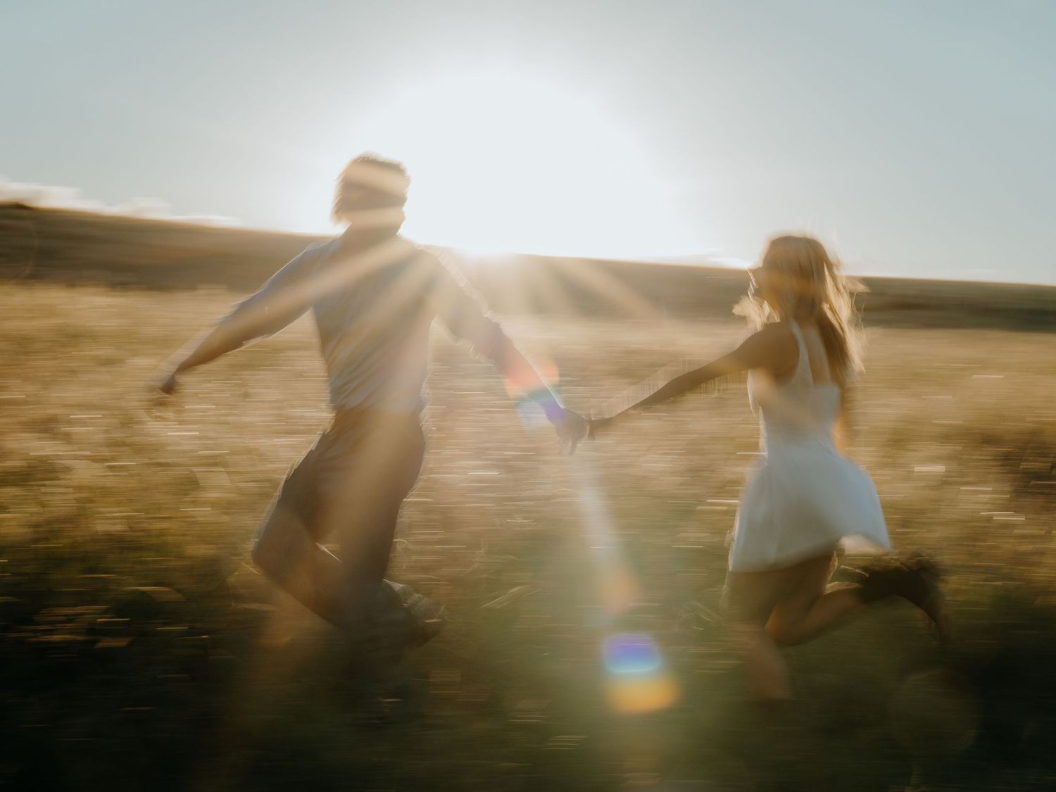 Couple holding hands running through golden sunlit field at sunset, romantic outdoor photography with lens flare.