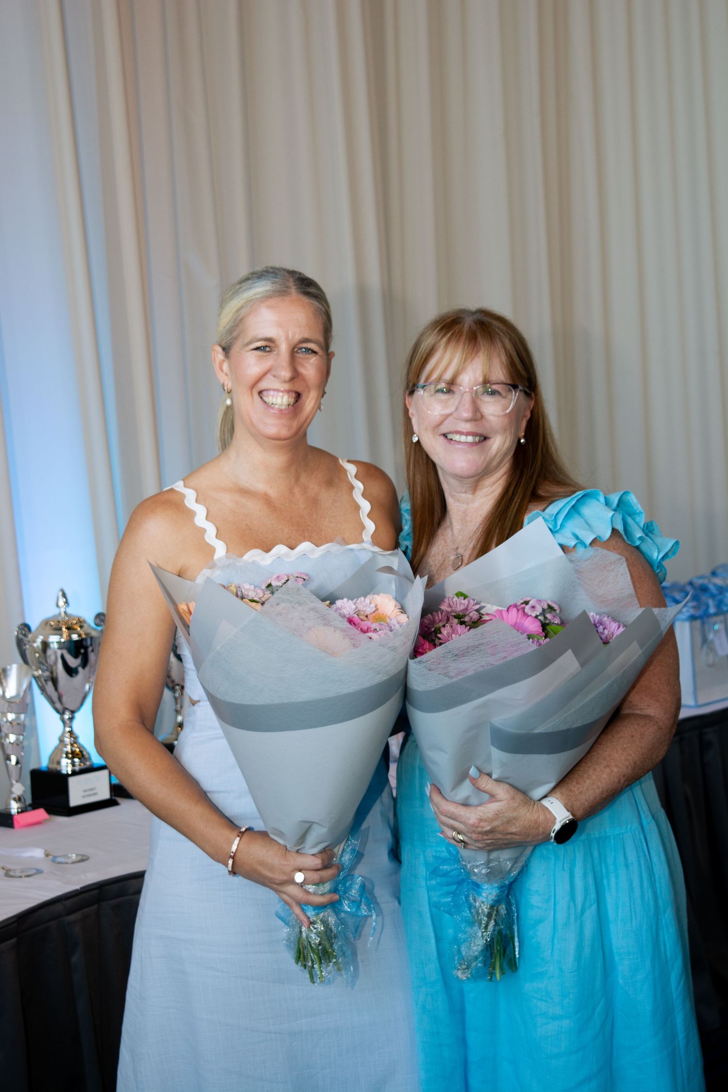 Two women holding flower bouquets pose together at an event.