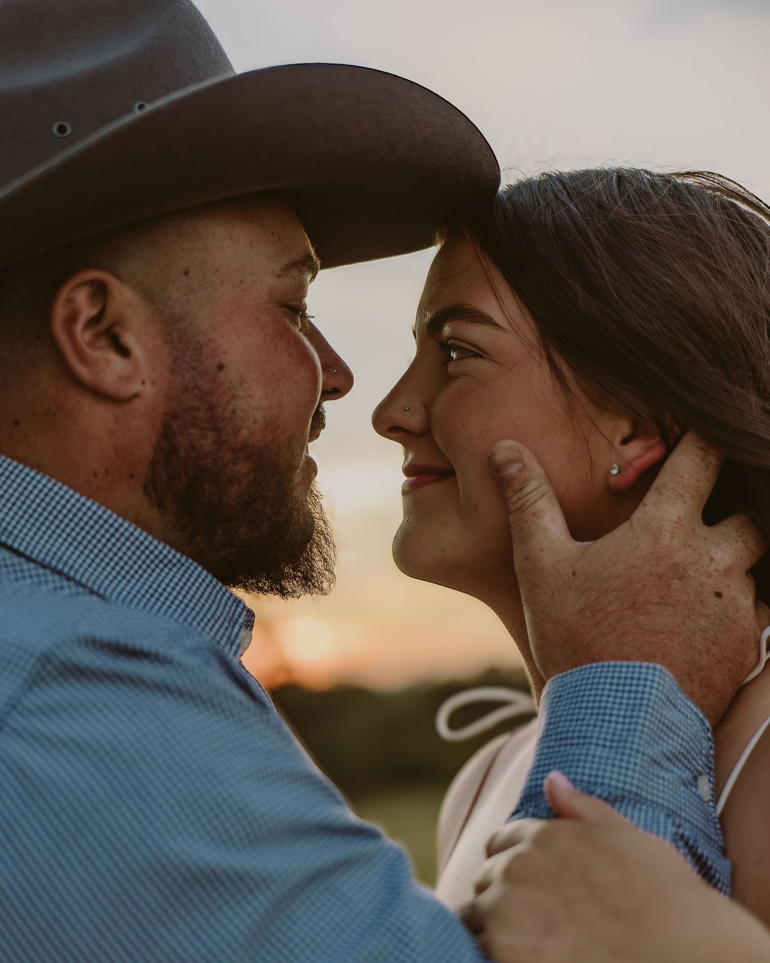 A bearded man in a cowboy hat tenderly cups a smiling woman's face during a romantic golden hour outdoor portrait.
