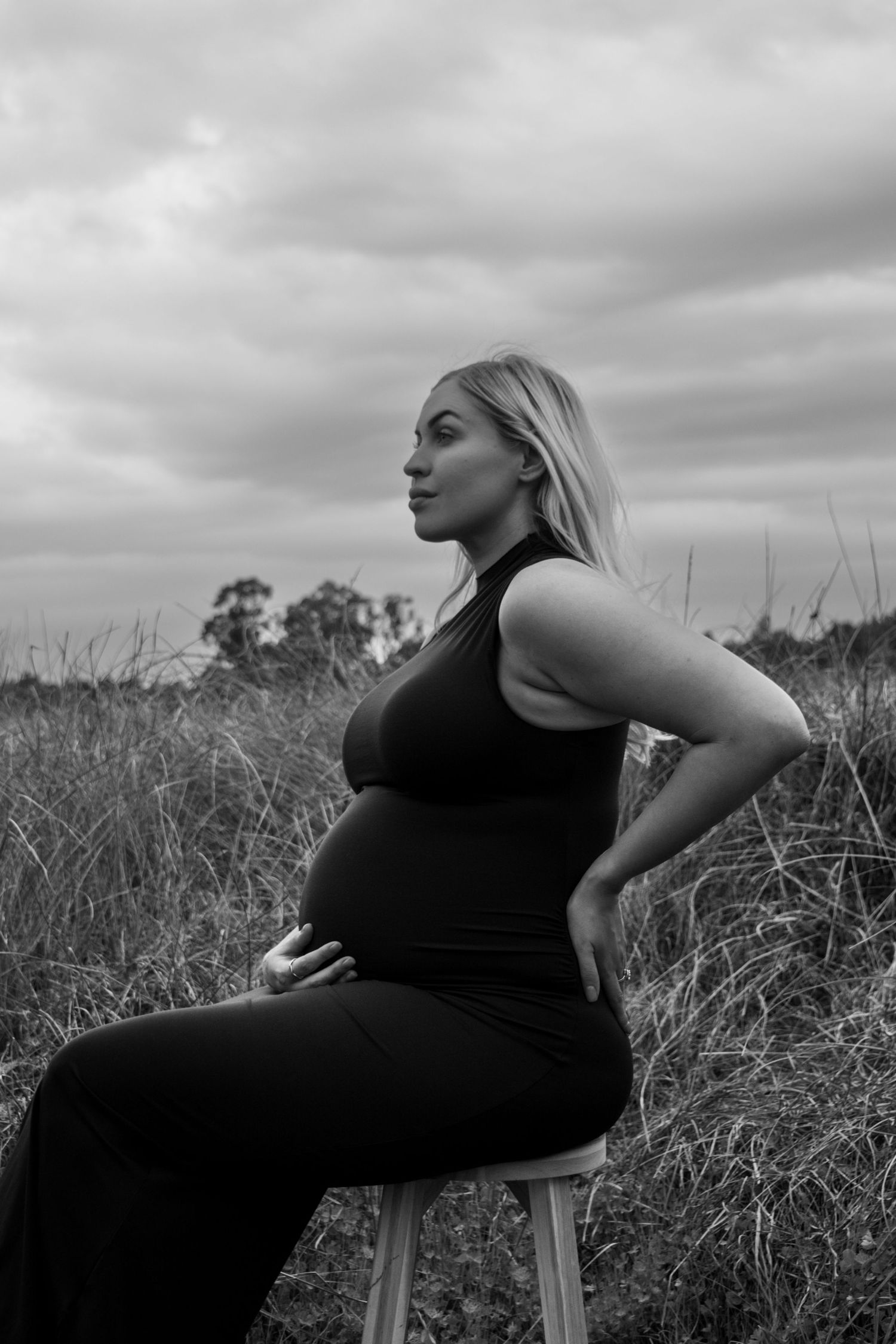 A black and white portrait of an expectant mother sitting on a stool in a grassy field under cloudy skies.