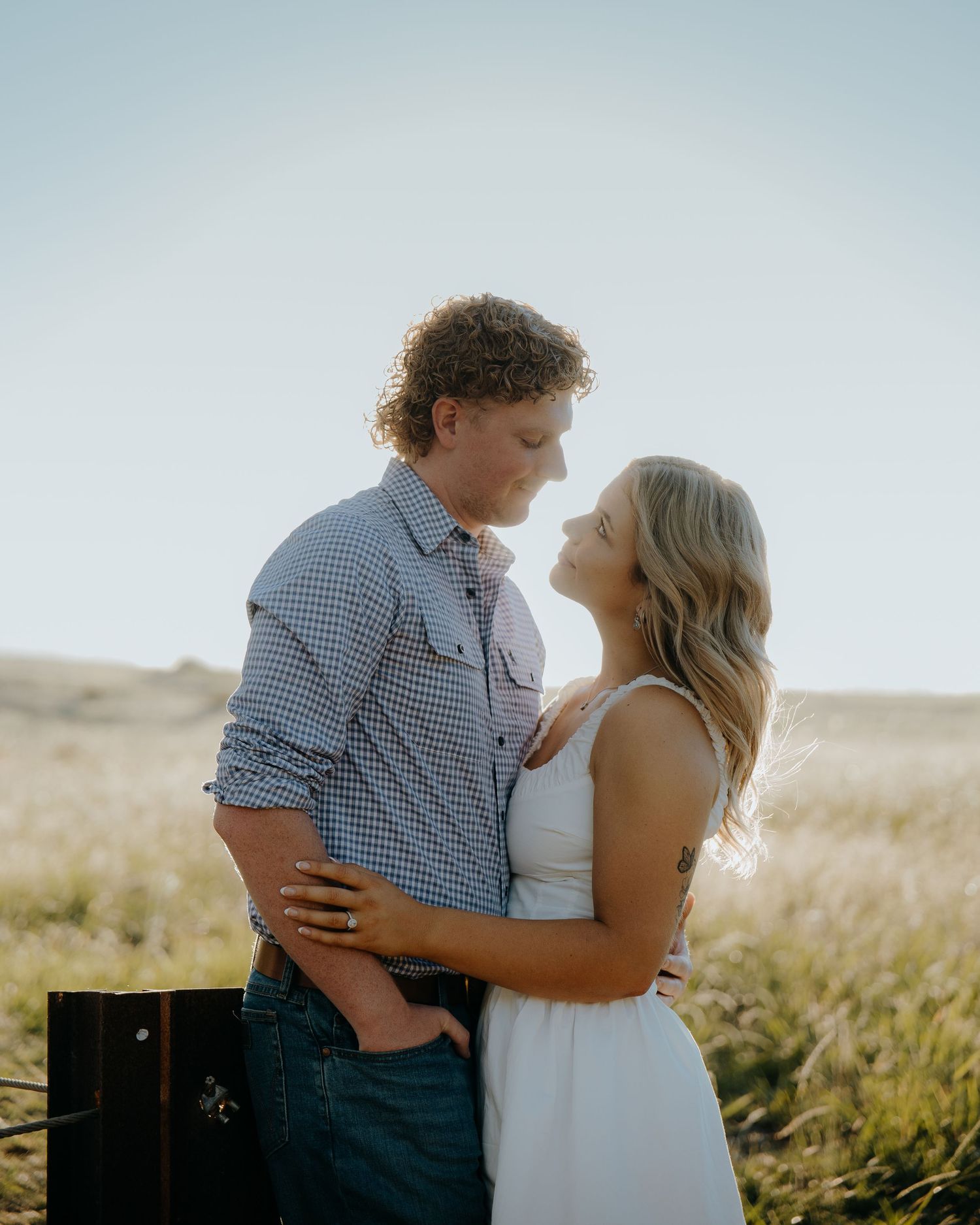 A couple shares a romantic moment in a golden field, the man in a blue shirt and woman in a white dress gazing at each other.