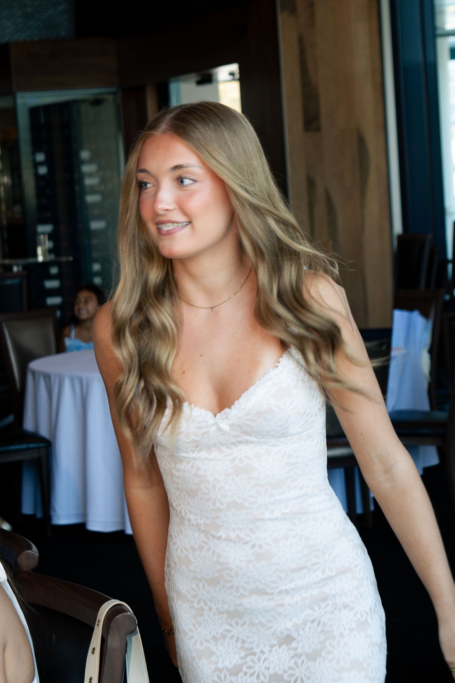 A woman in a white lace dress smiles while standing in an indoor venue.