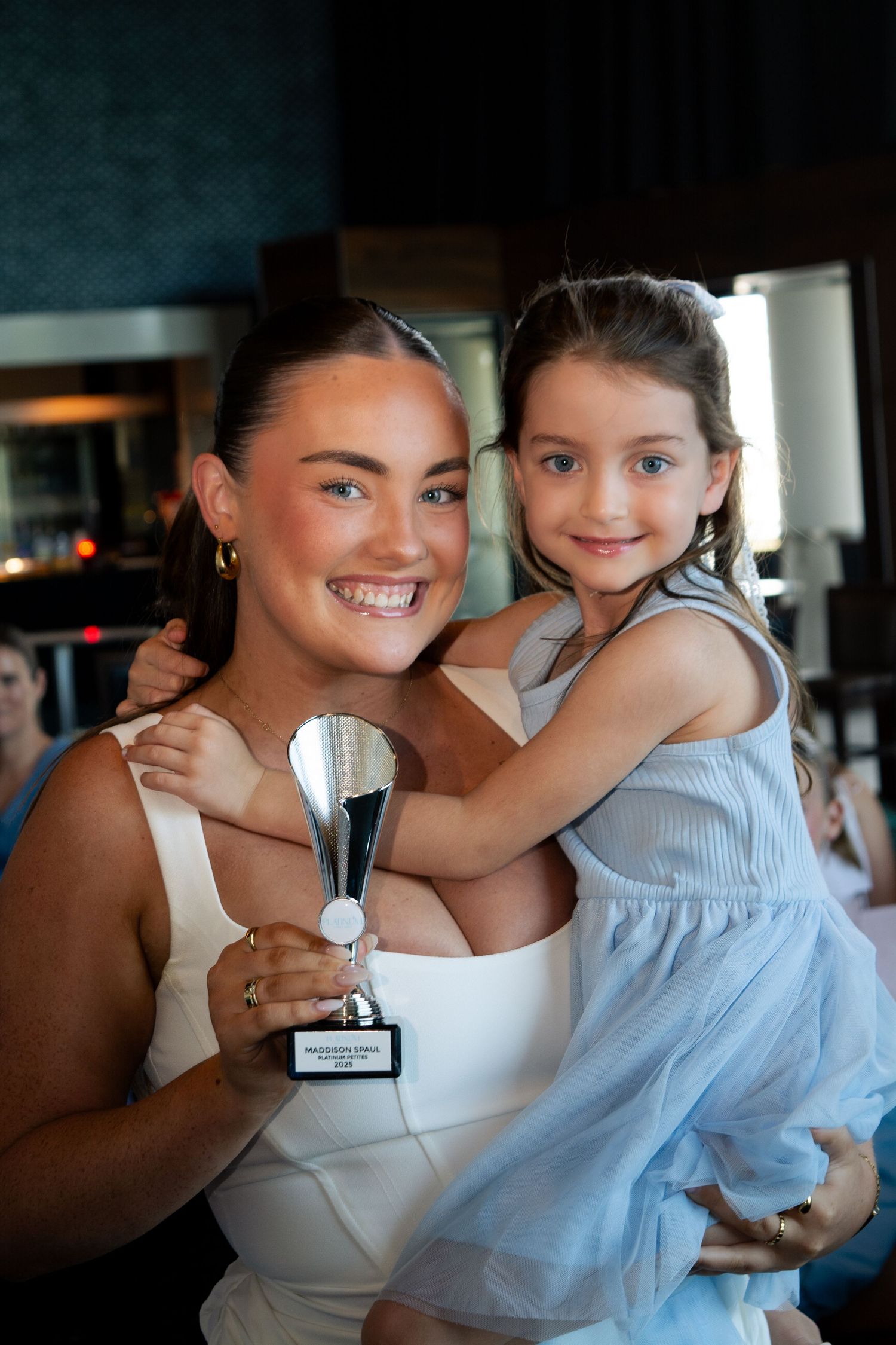 A person holds a trophy while posing with a young girl in a blue dress.