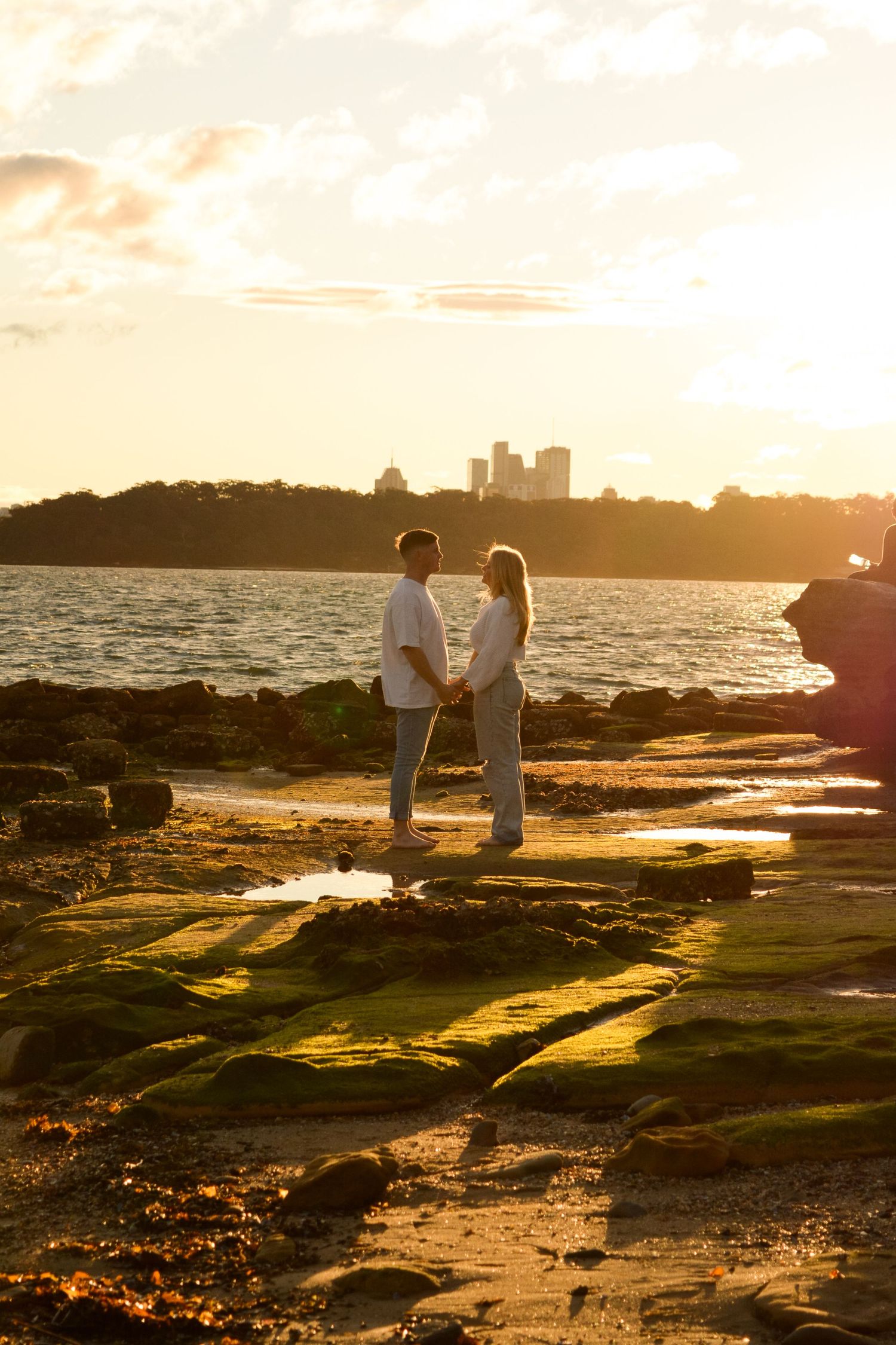 Silhouettes of two people sitting on rocky coastal terrain at sunset with city skyline in background.