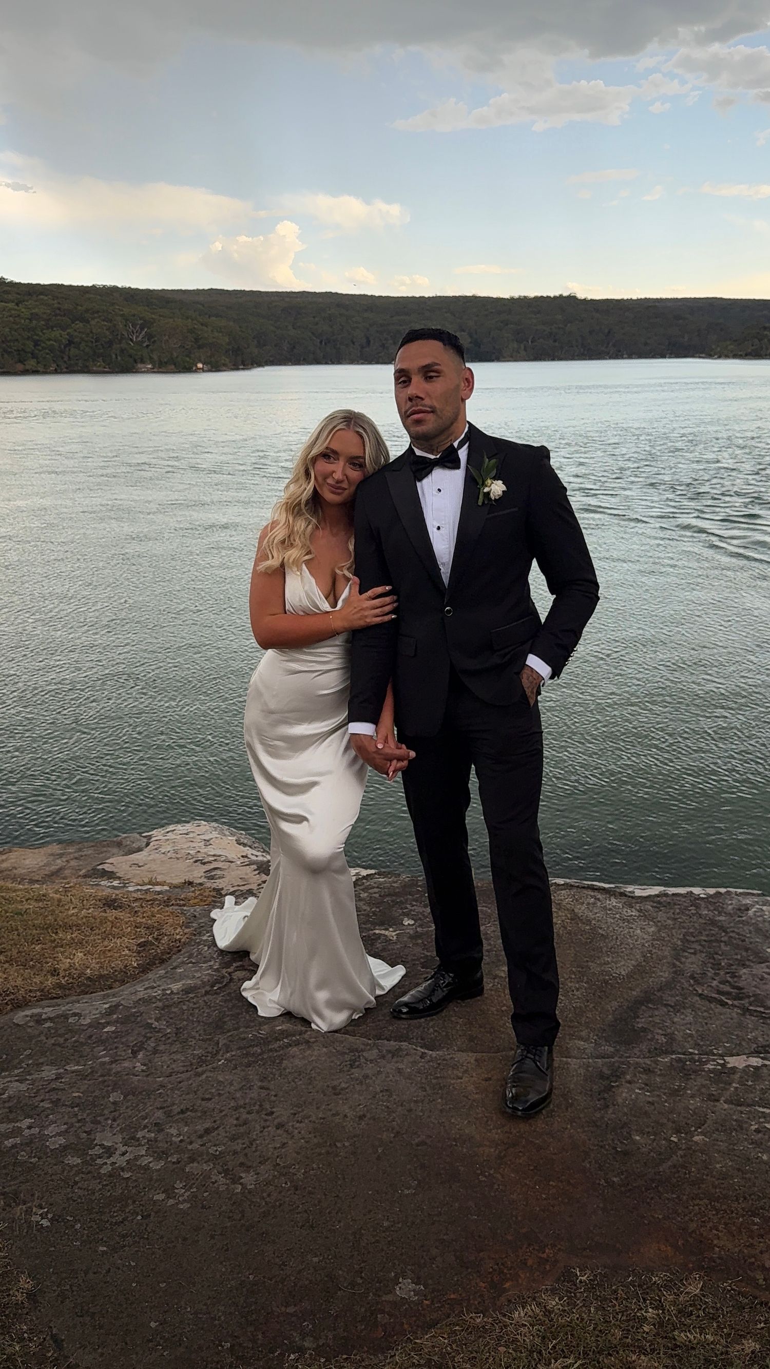 A wedding couple poses together on a rocky cliff overlooking a scenic waterfront at sunset.
