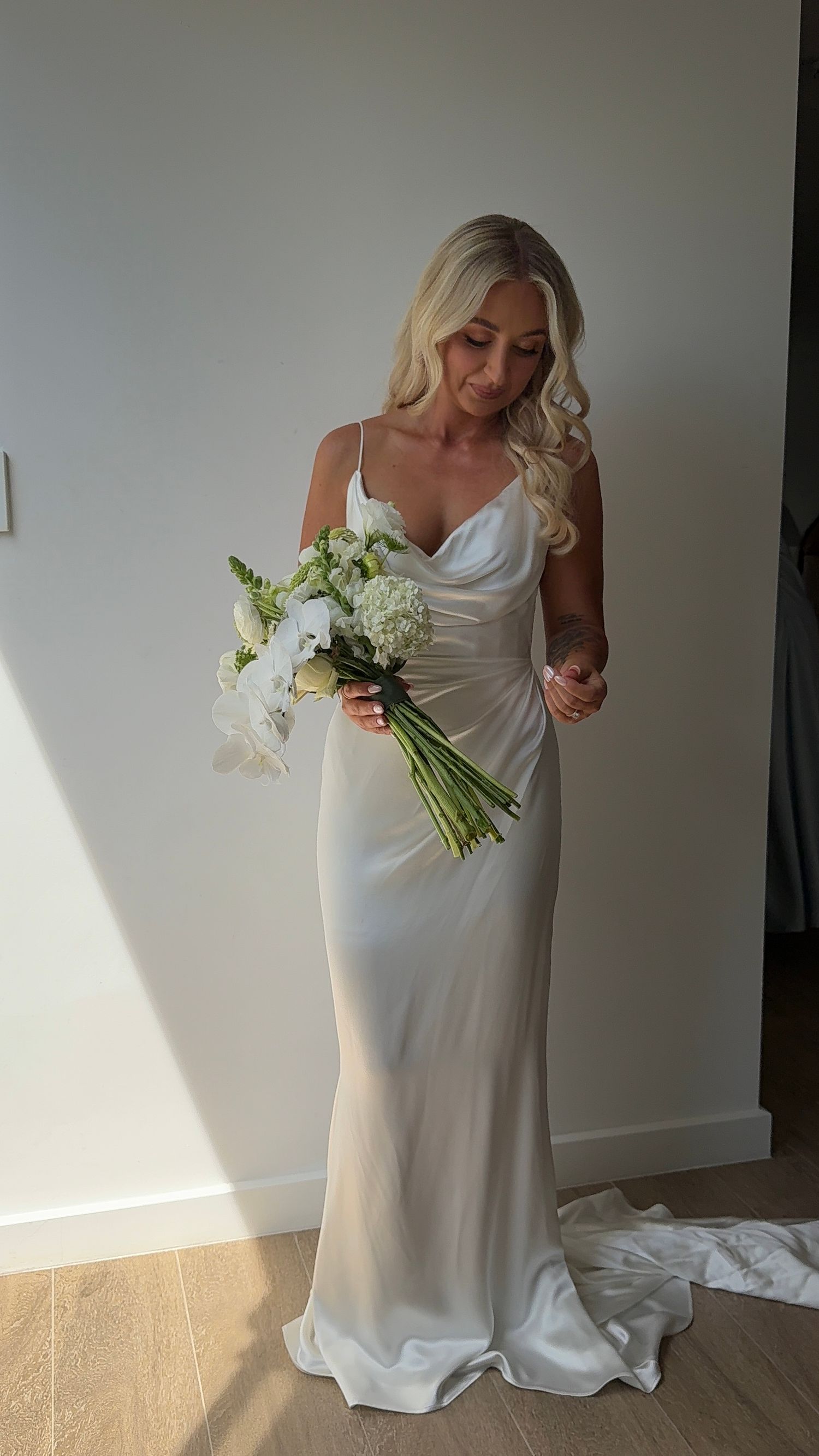 A bride in an elegant white gown holds a bouquet of white flowers while posing in natural window light.